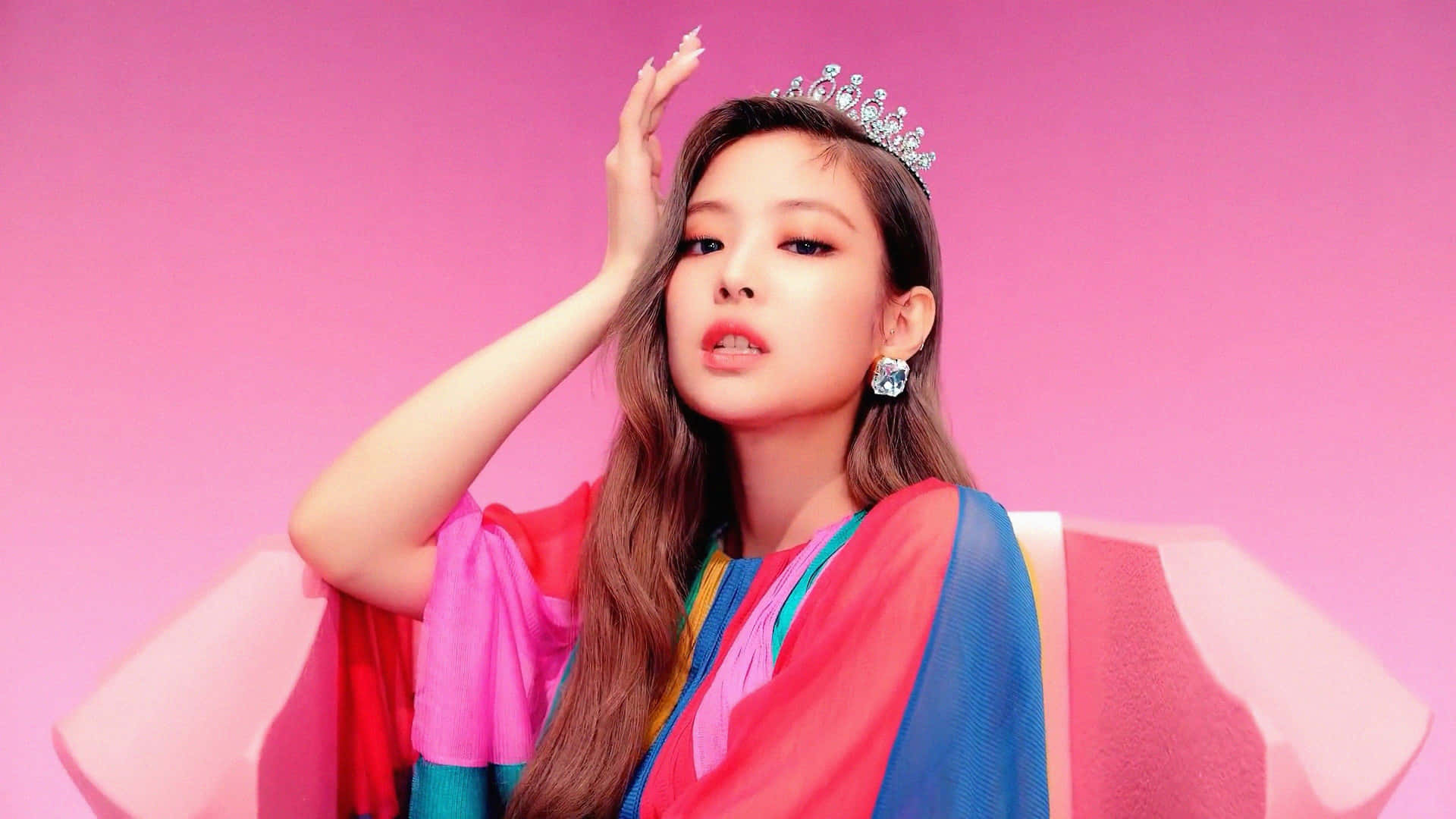 BLACKPINK Jennie Kim戴皇冠穿彩色禮服照片 壁紙 — 一位戴著閃亮頭飾、身穿色彩繽紛層次服裝的女性，在充滿活力的粉紅色背景前自信地擺姿勢。