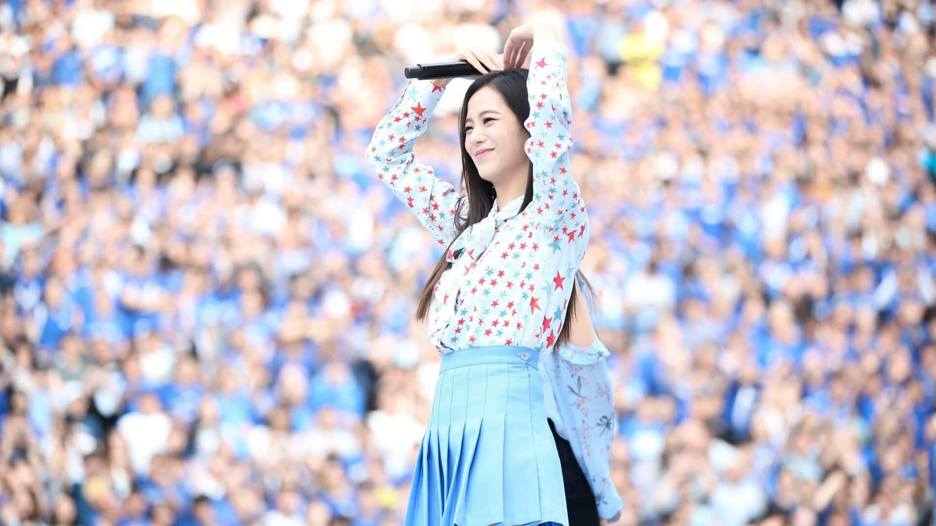 BLACKPINK Jisoo 機場愛心手勢 壁紙 — 一位微笑的表演者身穿星星圖案襯衫和藍色裙子，在藍衣粉絲的海洋前將麥克風高舉過頭頂。