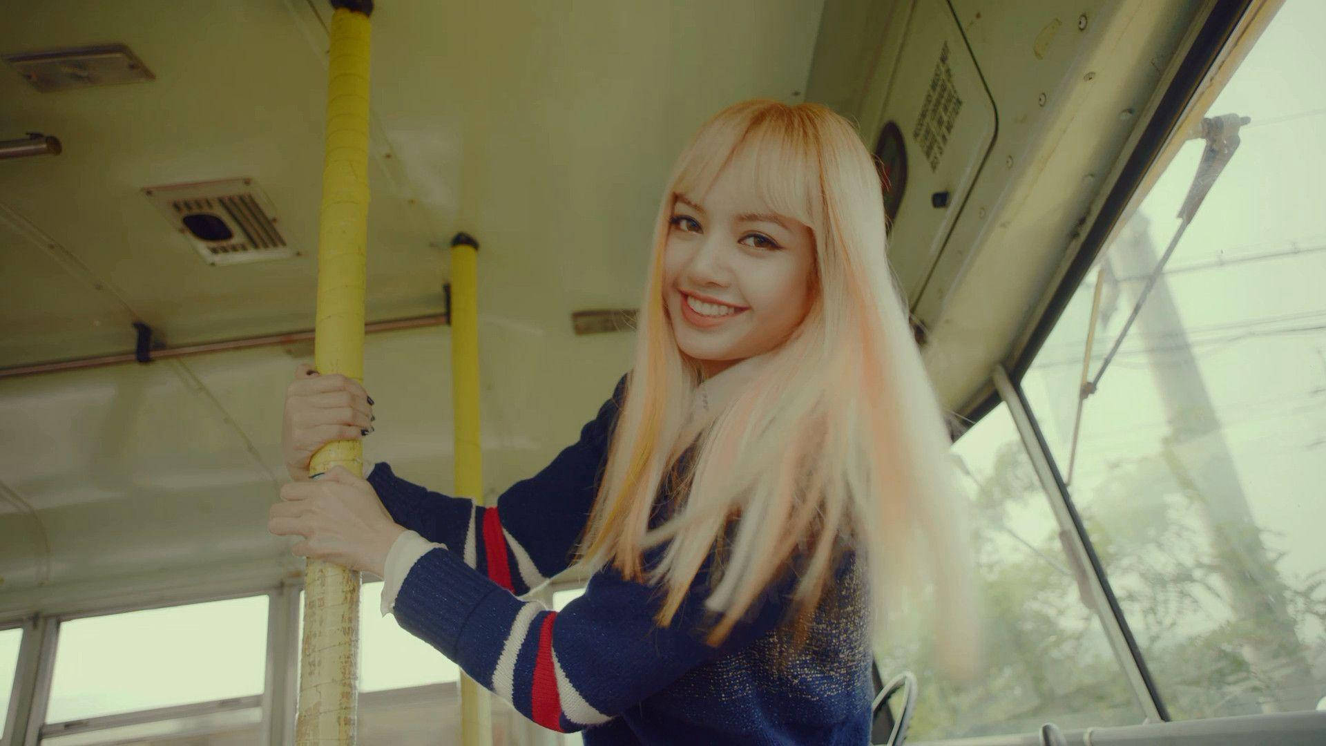 BLACKPINK LISA 乘巴士 壁紙 — 一位微笑的金髮年輕女子在柔和自然光下，於復古電車內握著一根黃色柱子。