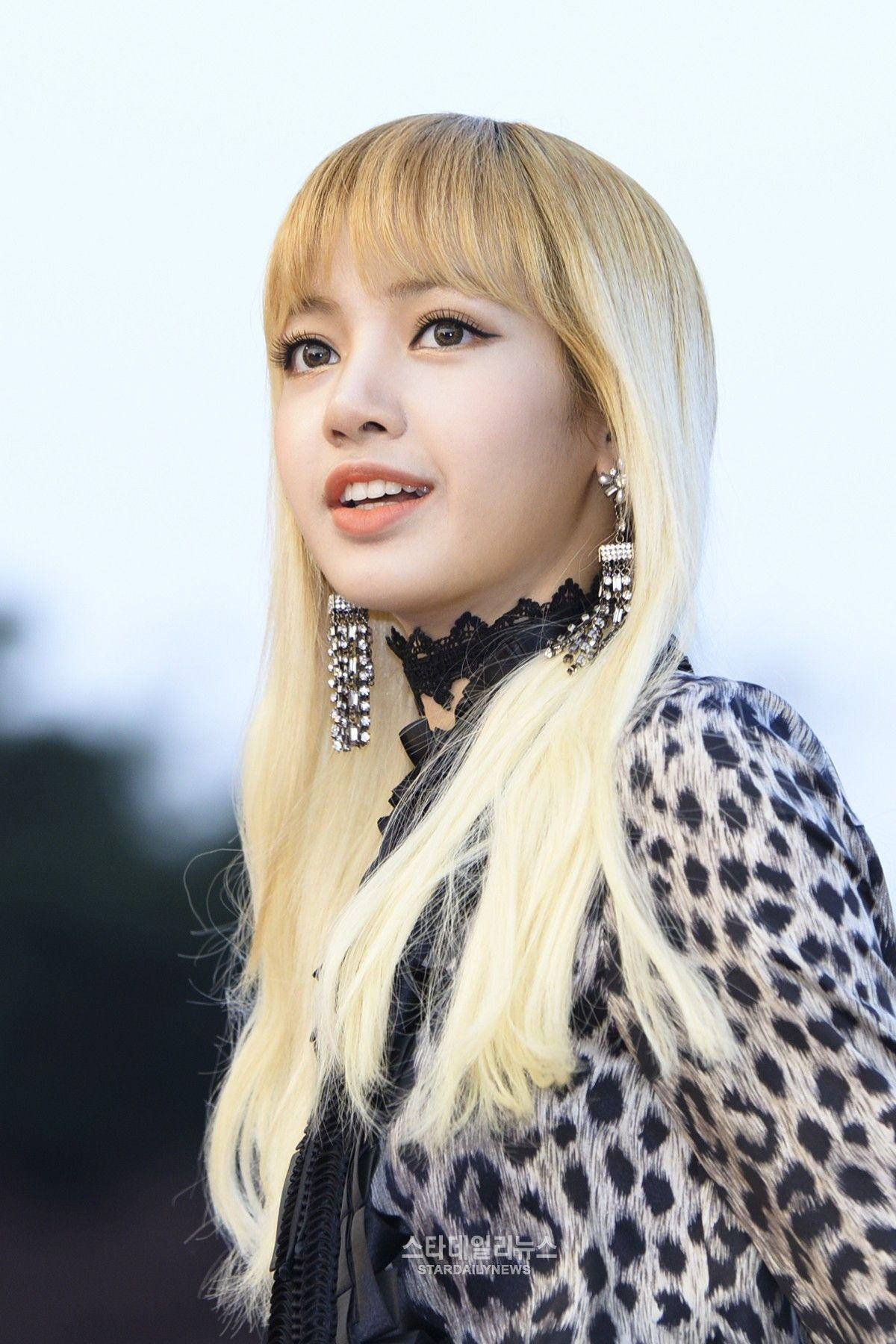 BLACKPINK LISA 動物紋造型 壁紙 — 一位年輕女性，長長的金色頭髮，大膽的妝容，穿著豹紋上衣和閃閃發光的吊燈耳環，背景柔和模糊。