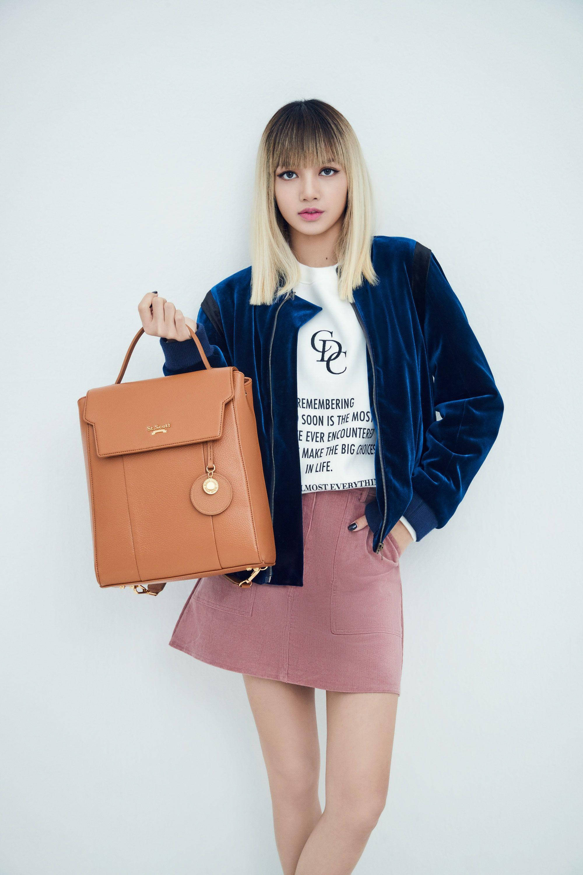 BLACKPINK LISA 手袋拍攝 壁紙 — 一位金髮年輕女性穿著藍色天鵝絨外套和粉紅色裙子，手持棕褐色皮革背包，背景為純白色。
