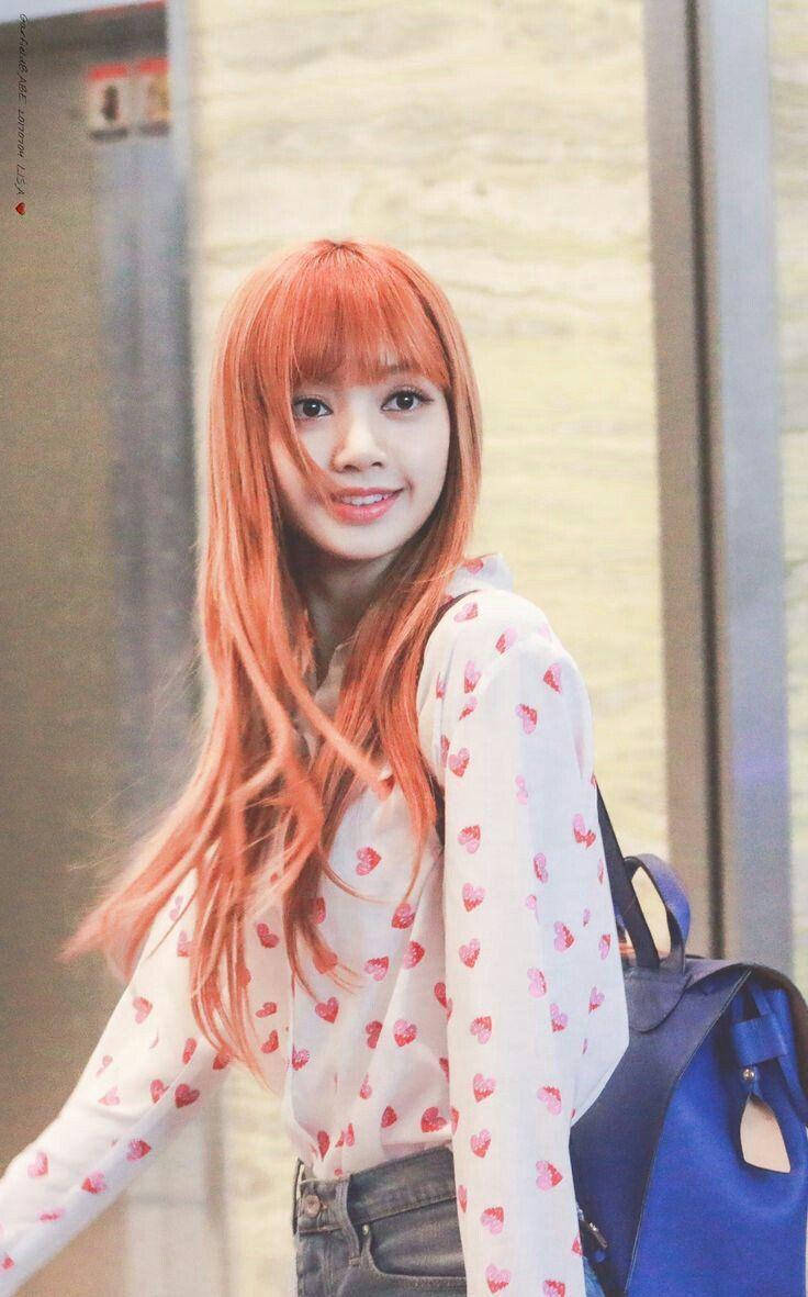 BLACKPINK LISA 真人隨拍 壁紙 — 一位擁有鮮豔橙色頭髮的年輕女性微笑著，穿著心形圖案襯衫，背著藍色背包。
