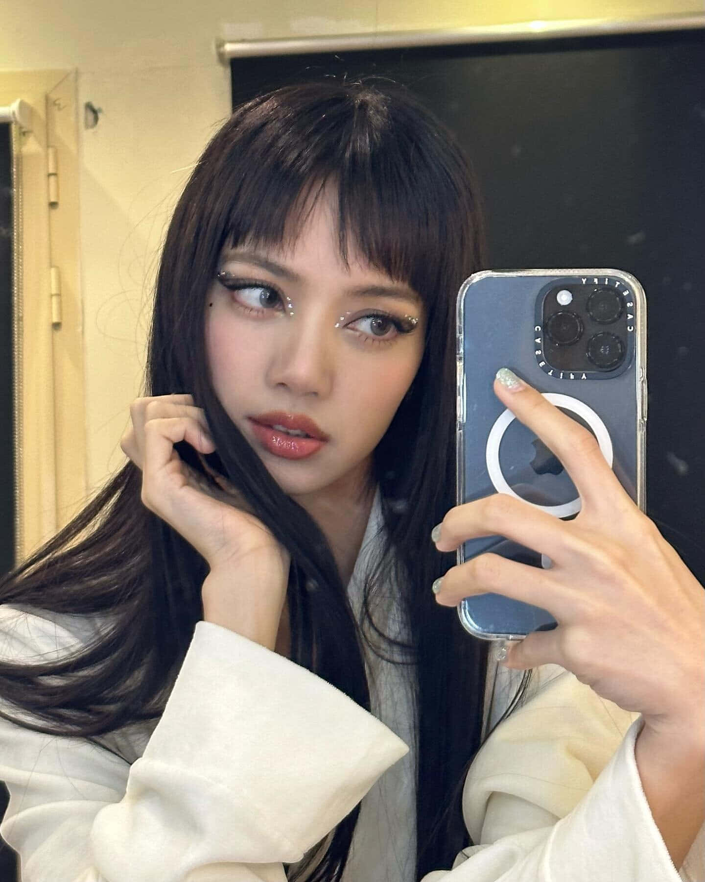BLACKPINK Lisa 自拍美照 壁紙 — 一位年輕女性，長黑髮，妝容大膽，在柔和光線的室內空間用智能手機拍鏡子自拍。