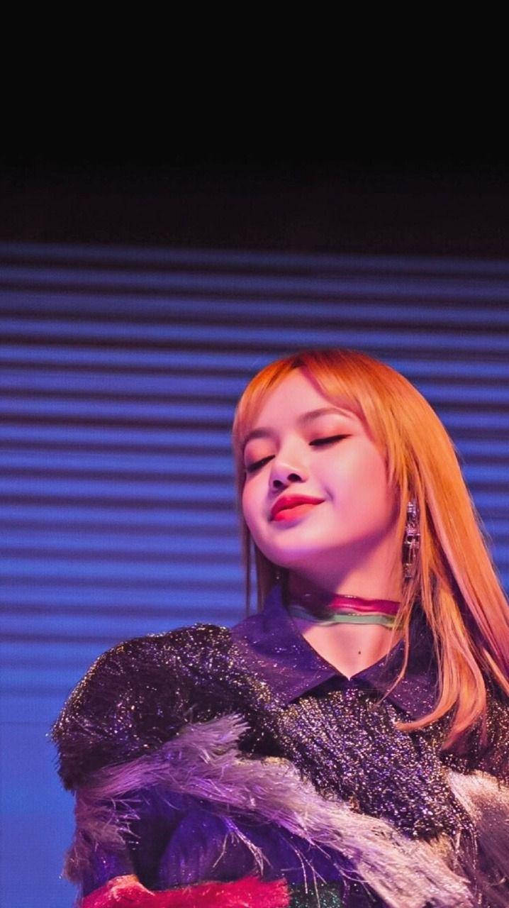 BLACKPINK LISA 閉眼微笑 壁紙 — 一位擁有橙色頭髮和閃亮服裝的表演者在充滿活力的藍色舞台燈光下溫柔地微笑。