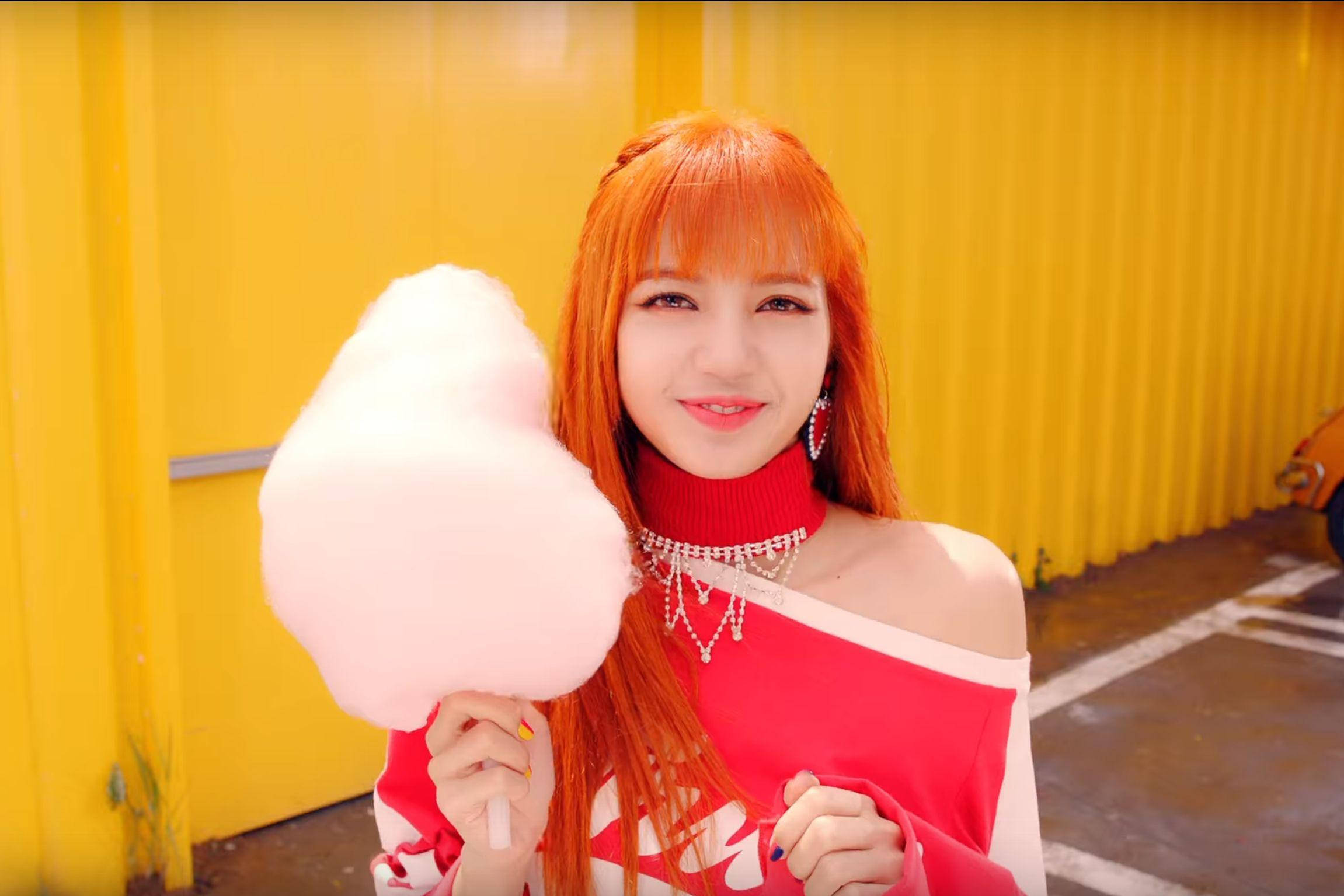 BLACKPINK LISA手持棉花糖 壁紙 — 一位開朗的女士，擁有火紅色的頭髮，在鮮黃色的波紋牆前拿著一個巨大的粉紅色棉花糖。