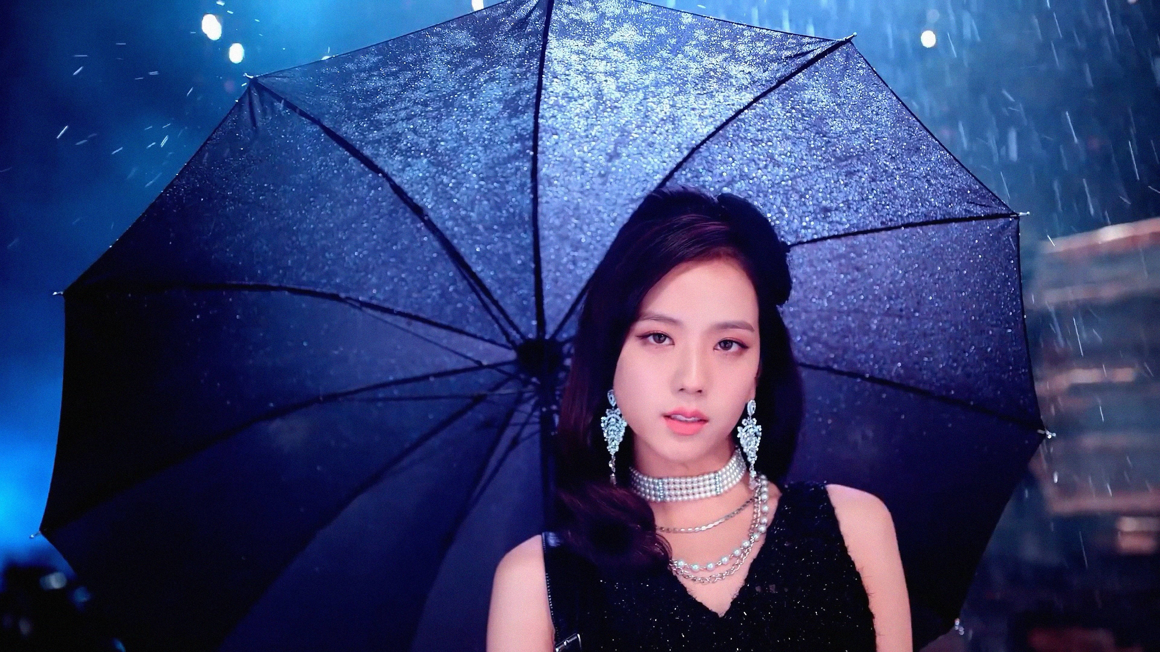 BLACKPINK 成員 Jisoo 持傘雨中電腦桌面壁紙 壁紙 — 一位深色頭髮、佩戴閃亮珠寶的女性，在藍色燈光的城市背景下，於落雨中手持一把閃閃發光的黑色雨傘。