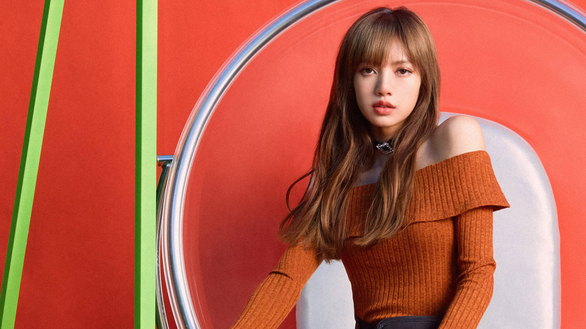 BLACKPINK 成員 Lisa 穿橙色上衣電腦桌面壁紙 壁紙 — 一位長髮棕色的年輕女性坐在現代椅子上，背景是大膽的紅色和綠色幾何圖案。
