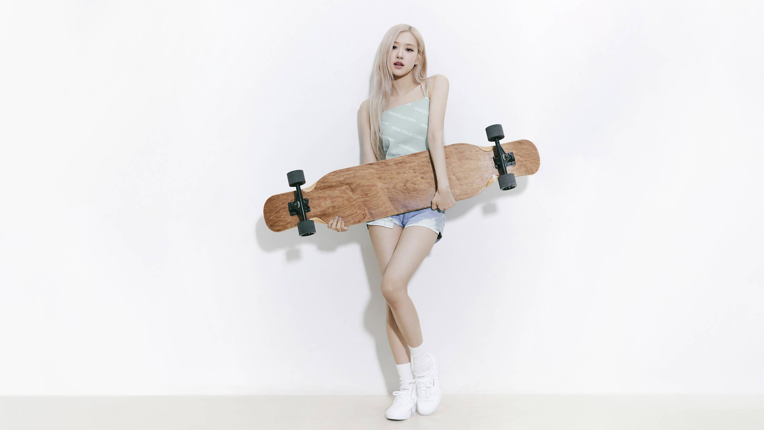 BLACKPINK 成員 Rosé 持滑板電腦桌面壁紙 壁紙 — 一位長金髮的年輕女性在簡約的工作室環境中，將木製長板靠在純白牆上。
