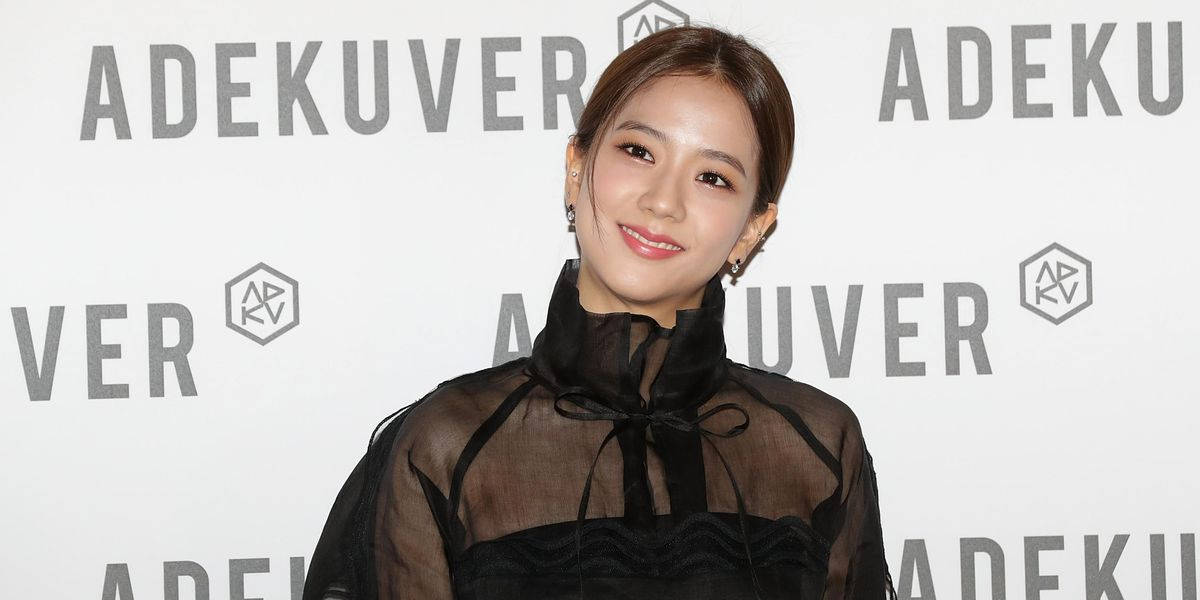 BLACKPINK 智秀 Adekuver 活動照 壁紙 — 一位微笑的女性穿著黑色透視襯衫，領口有蝴蝶結，站在品牌背景前。