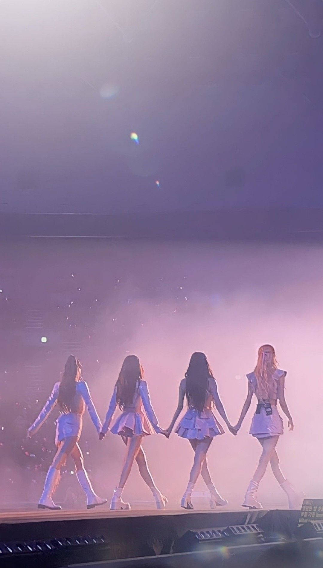 BLACKPINK 舞台美學壁紙 壁紙 — 四位女表演者手牽手走過一個煙霧瀰漫、沐浴在紫色光線和鏡頭光暈中的舞台。