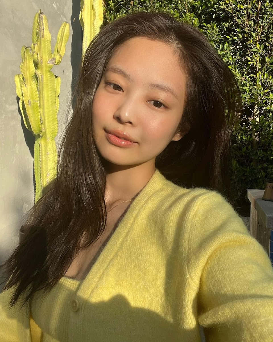 BLACKPINK成員Jennie身穿黃色針織開衫照片 壁紙 — 一位身穿柔軟黃色開襟毛衣的年輕女性，在陽光照射的牆壁旁，對著一株鮮綠色的仙人掌溫柔微笑。