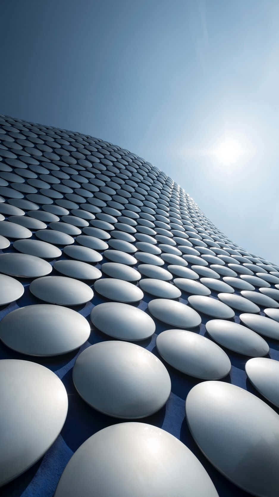 現代建築：伯明翰牛環區Blobitecture 壁紙 — 在明亮的藍天下，一座未來主義建築的低角度視圖，其彎曲的外牆由金屬銀色圓盤構成。