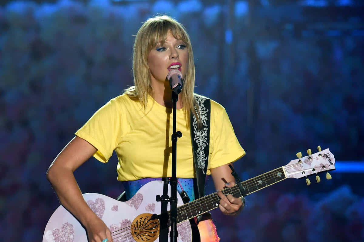 Taylor Swift Lover 桌布 壁紙 — 一位金髮女子身穿黃色襯衫，在藍色舞台燈光下，一邊彈奏心形圖案的原聲結他，一邊對著咪高峰唱歌。