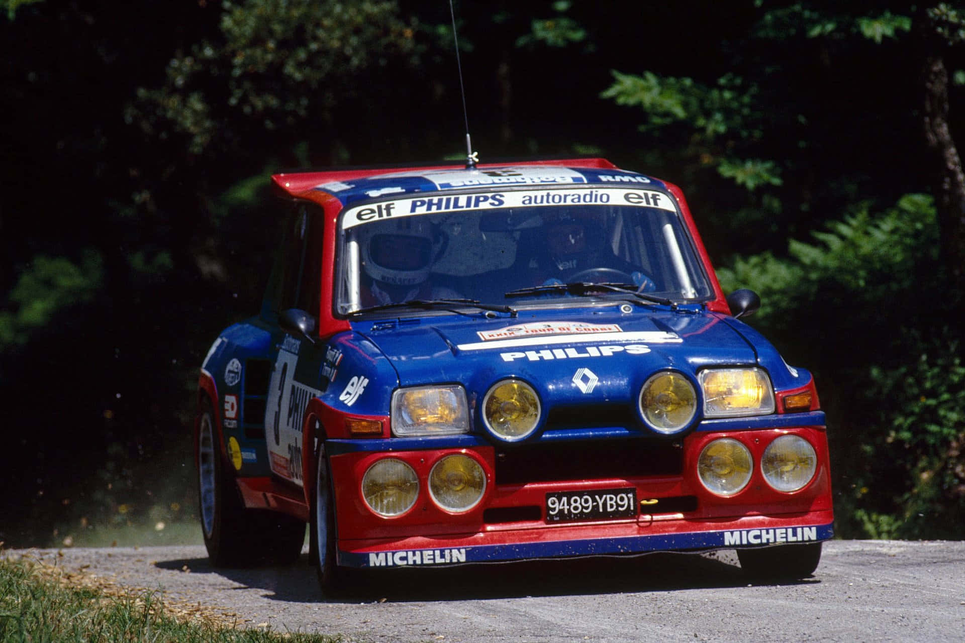 Renault 5 Turbo 桌布 壁紙 — 一輛藍色和紅色的雷諾5 Turbo拉力賽車在明亮的陽光下，在森林道路上疾馳，揚起灰塵。