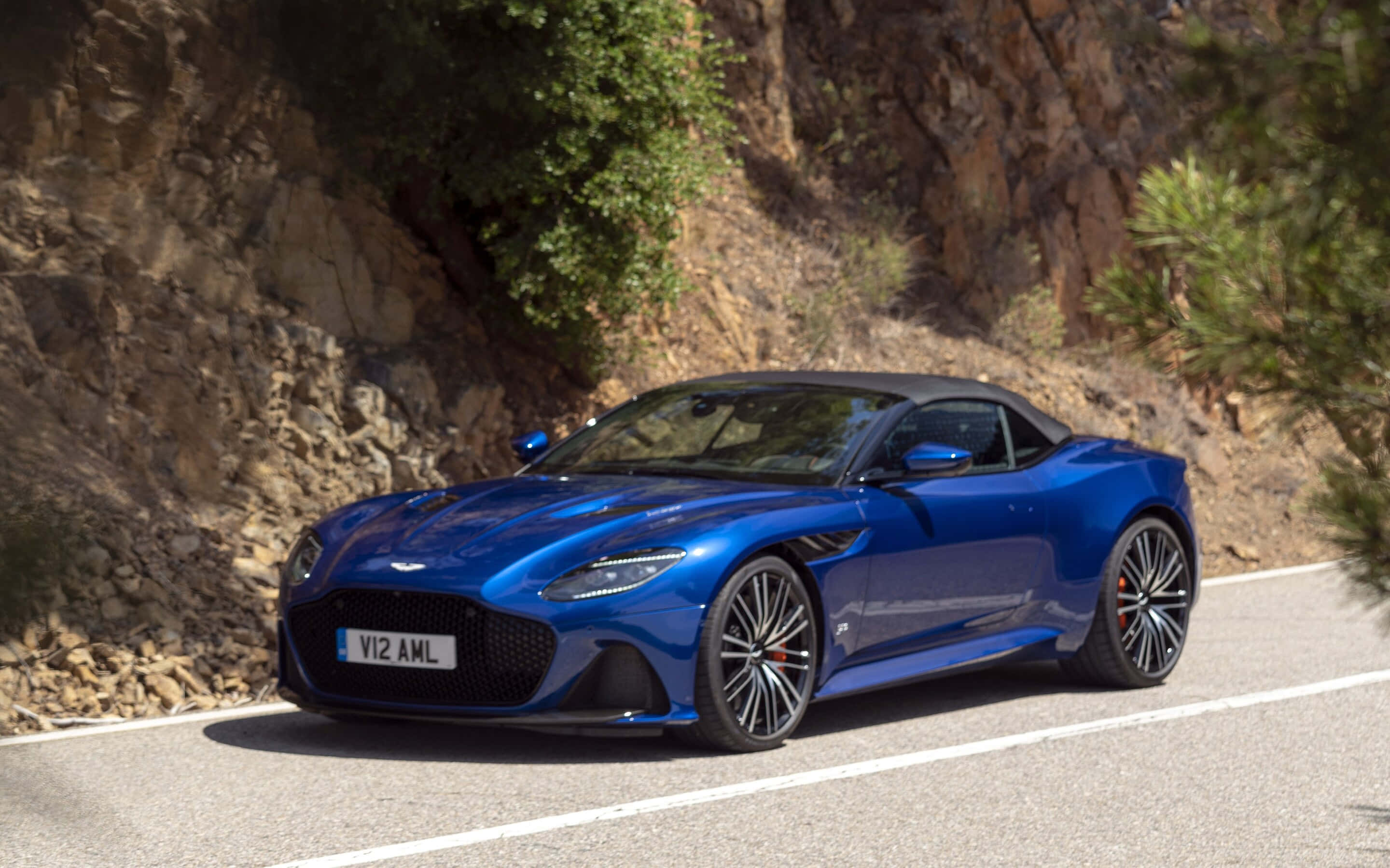 Aston Martin Dbs Superleggera 桌布 壁紙 — 一輛充滿活力的藍色 Aston Martin V12 Vantage 開篷跑車沿著蜿蜒的山路巡航，兩旁是岩石峭壁和綠意。
