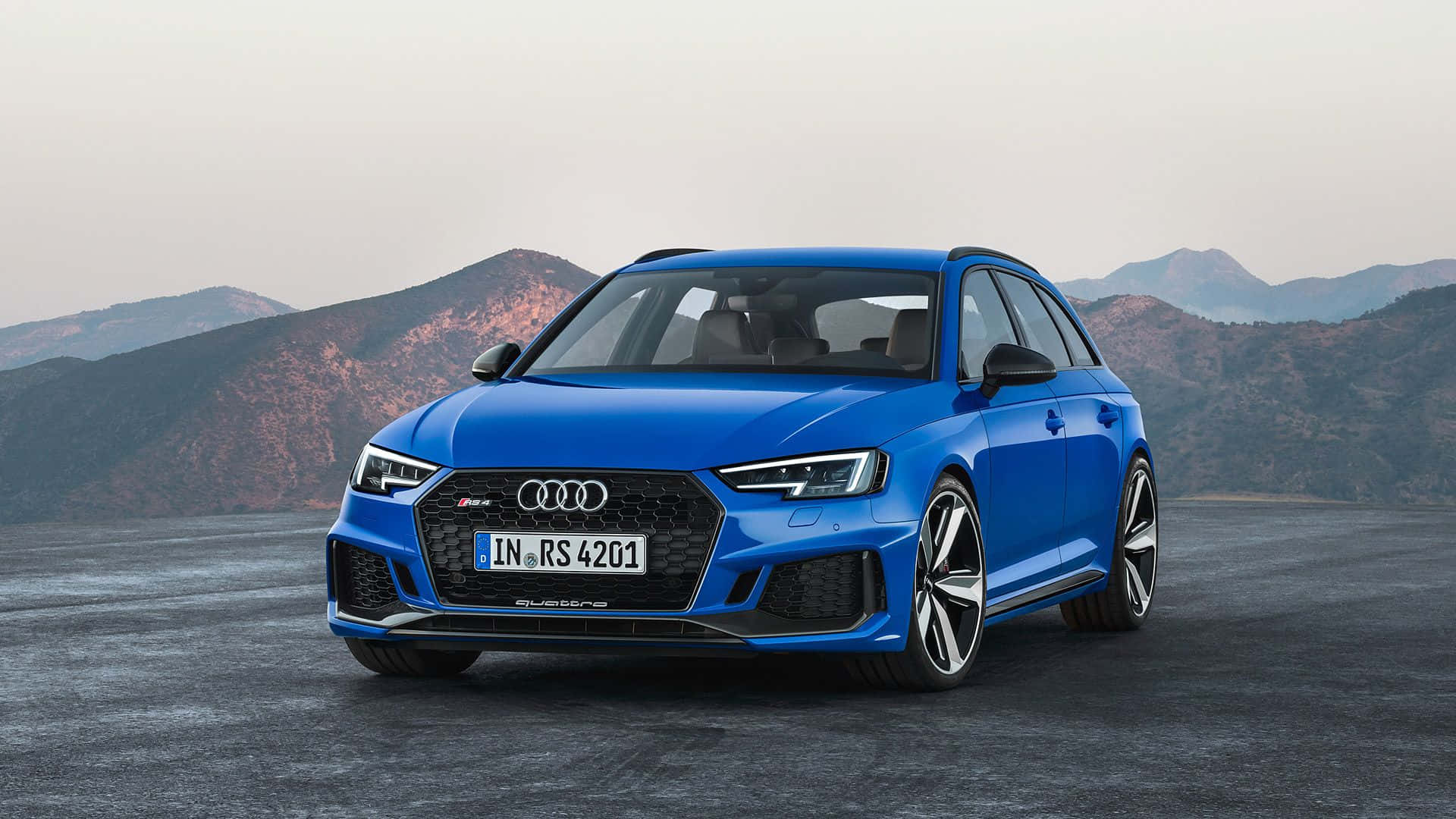 Audi Rs4 桌布 壁紙 — 一輛充滿活力的藍色 Audi RS4 Avant 運動旅行車停在鋪砌的路面上，背景是朦朧的崎嶇山脈。