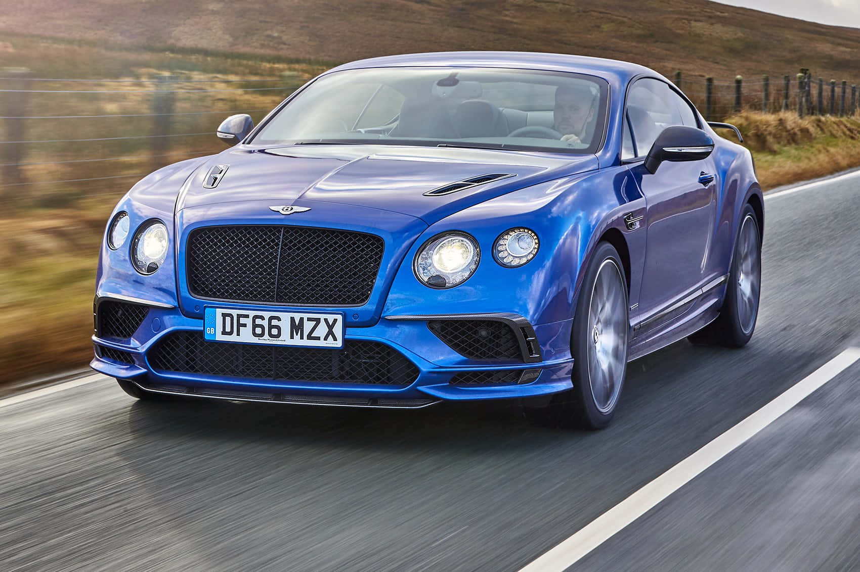 Bentley Continental Supersports 桌布 壁紙 — 一輛充滿活力的藍色賓利 Continental GT 在蜿蜒的鄉村道路上飛馳，背景是模糊的金色草丘。