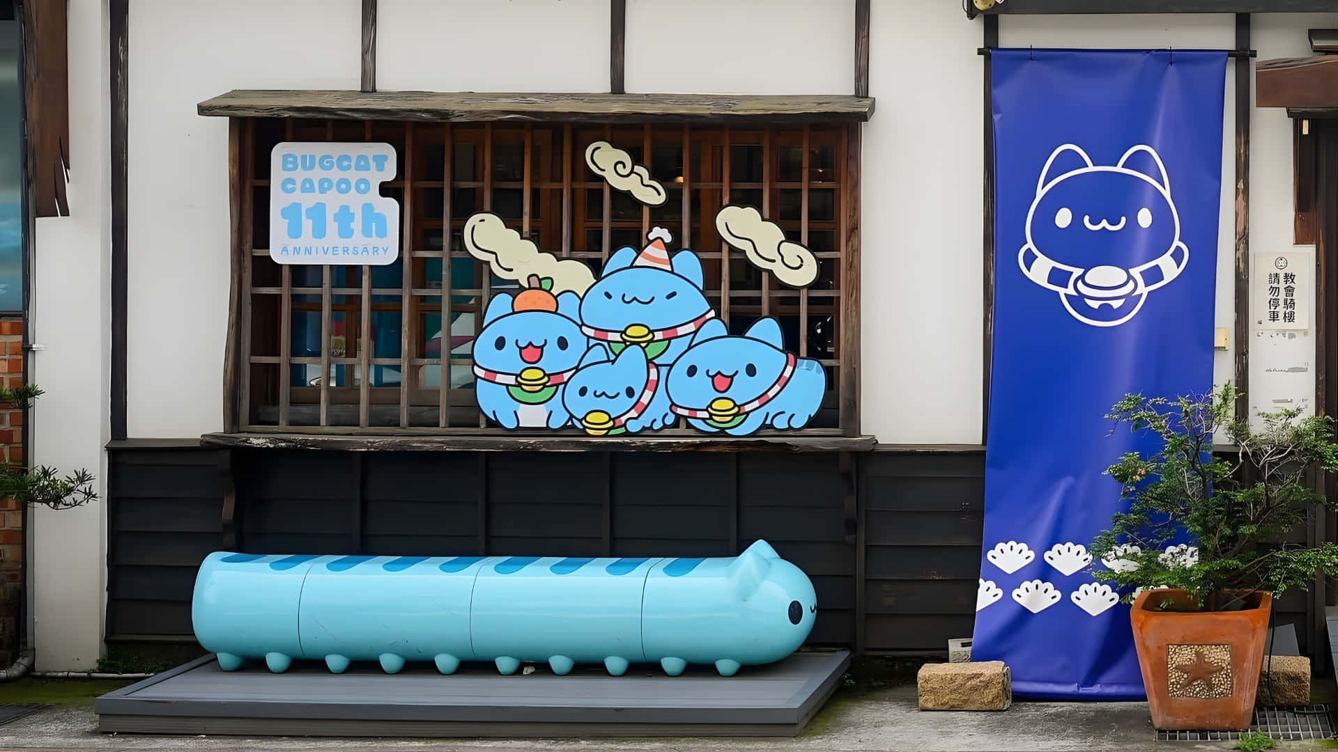  壁紙 — 藍色卡通毛毛蟲角色在傳統日本建築外牆上慶祝11週年，配有相配的藍色橫幅和大型毛毛蟲雕塑。