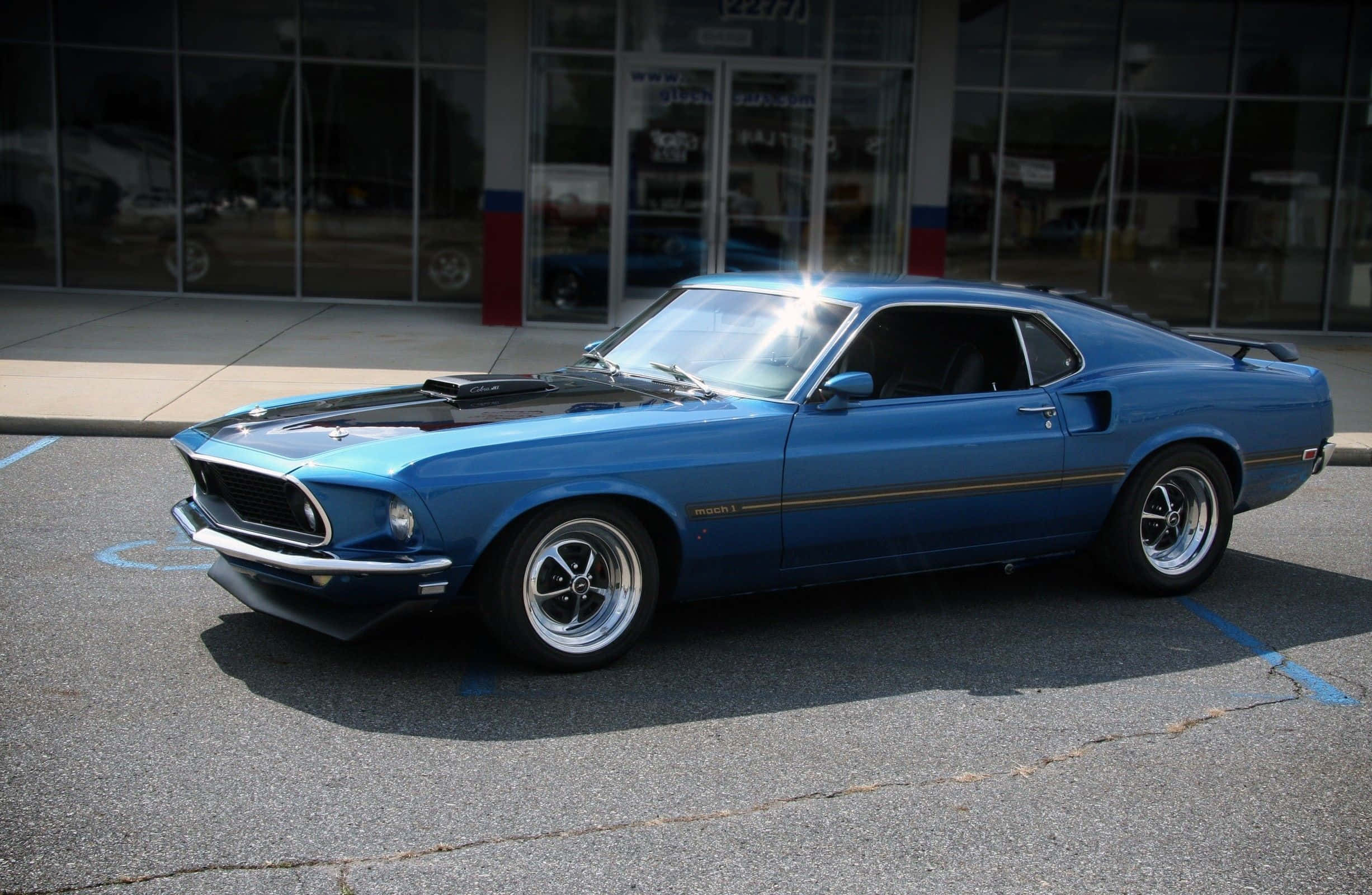 Ford Mustang Mach 1 桌布 壁紙 — 一輛充滿活力的藍色經典肌肉車，帶有黑色引擎蓋條紋，在明亮的陽光下閃閃發光，停在玻璃幕牆建築前。