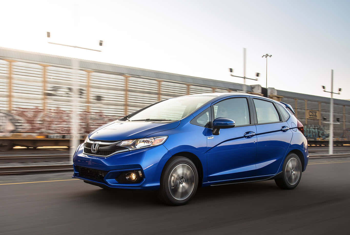Honda Fit 桌布 壁紙 — 一輛充滿活力的藍色本田Fit混合動力車在城市道路上疾馳，背景有動態模糊和塗鴉覆蓋的火車軌道。