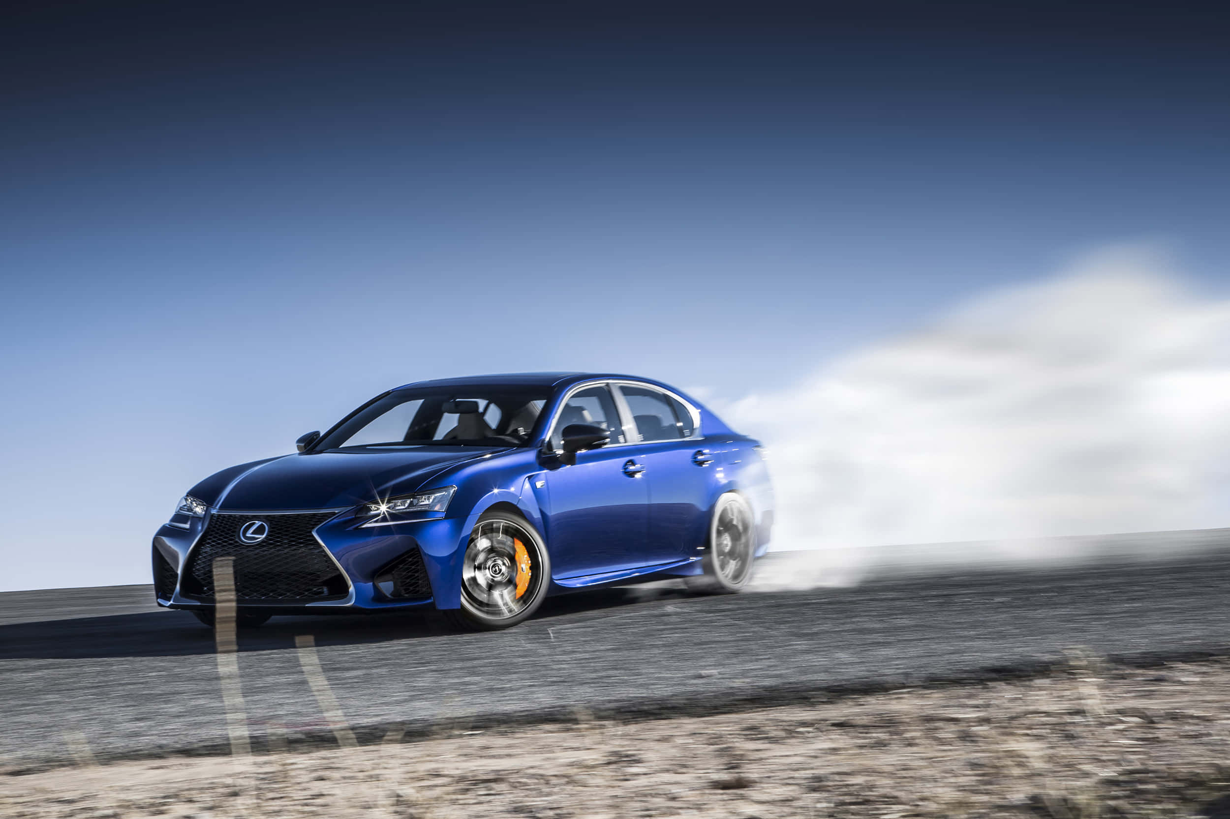 Lexus Gs F 桌布 壁紙 — 一輛充滿活力的藍色Lexus運動轎車在晴朗天空下的沙漠道路上飄移，後輪冒出滾滾濃煙。