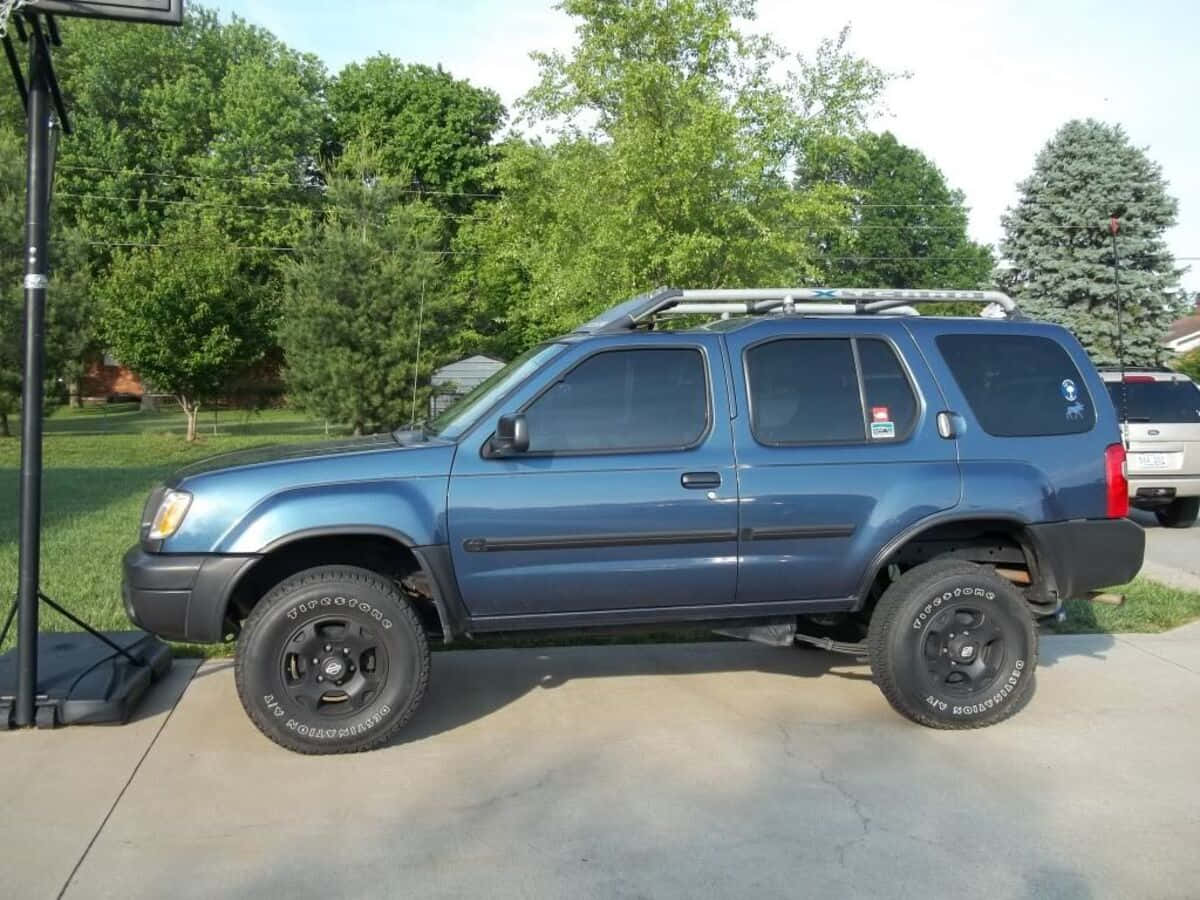 Nissan Xterra 桌布 壁紙 — 一輛升高底盤的藍色SUV，配備黑色輪圈和車頂行李架，停泊在混凝土車道上，周圍環繞著綠樹。