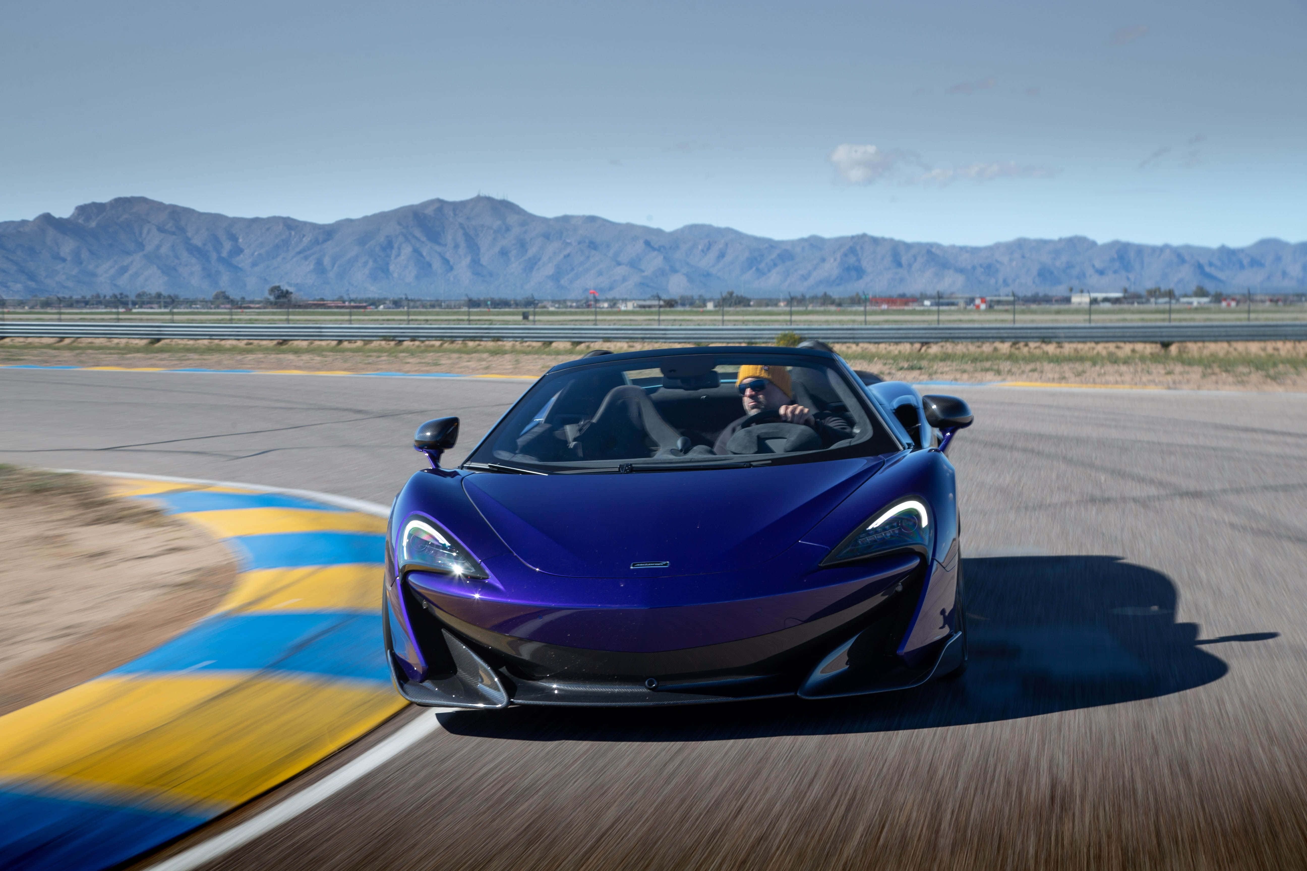 Mclaren 600lt Spider 桌布 壁紙 — 一輛充滿活力的藍色McLaren跑車在晴朗天空下，以群山為背景，在賽車場彎道上飛馳。