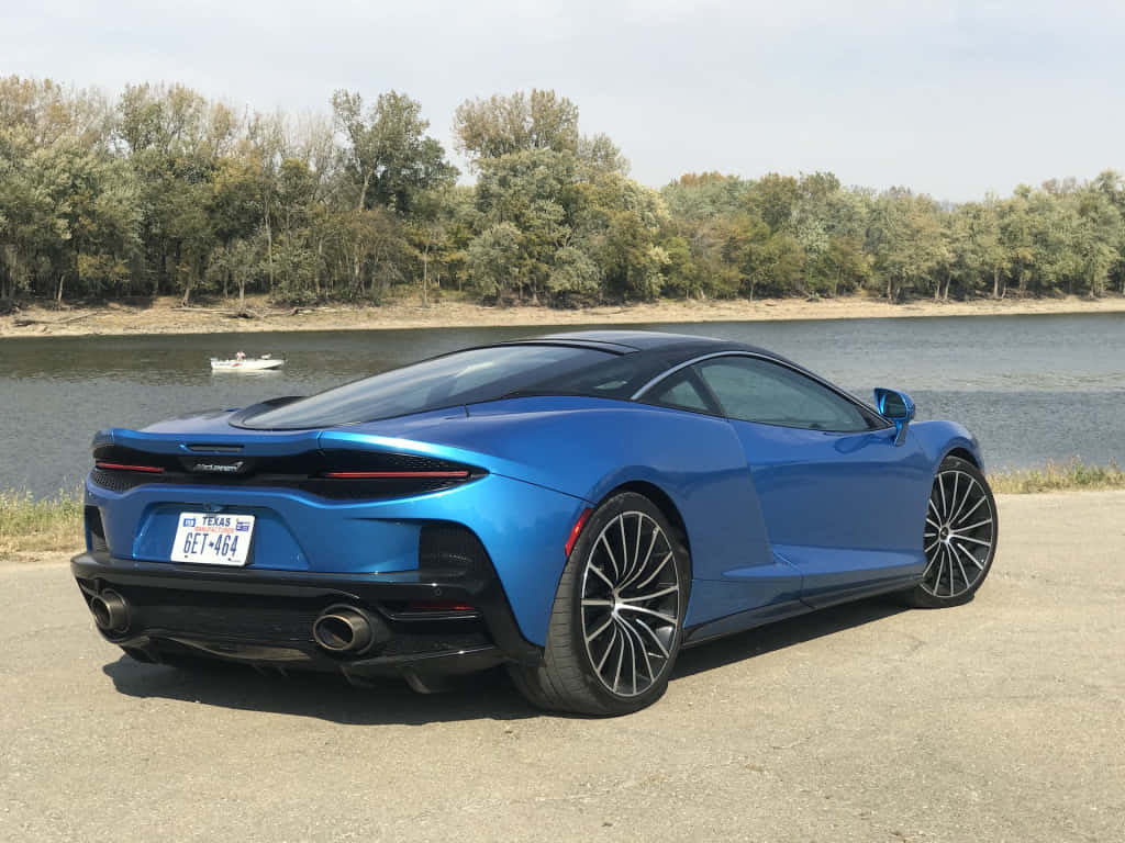 Mclaren Gt 桌布 壁紙 — 一輛鮮藍色McLaren跑車停泊在河邊，背景有樹木和小船。