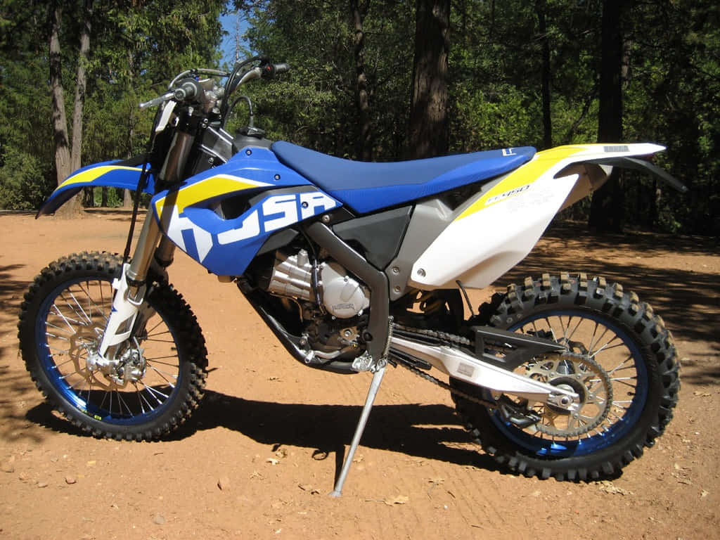  壁紙 — 一架藍色、黃色和白色的 Husqvarna 越野電單車停泊在陽光普照的泥徑上，周圍是高聳的樹木。