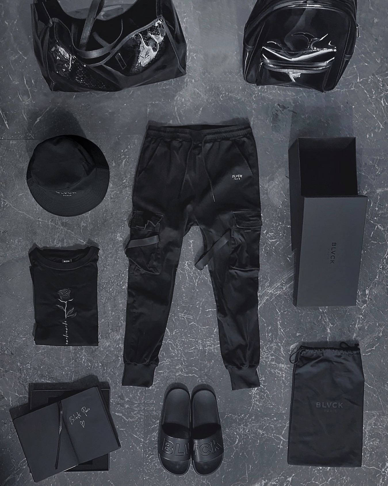 Blvck Paris 全黑服裝平鋪 壁紙 — 一個極簡主義的黑色時尚平鋪圖，展示BLVCK品牌的服裝配飾和手袋，排列在灰色大理石表面上。
