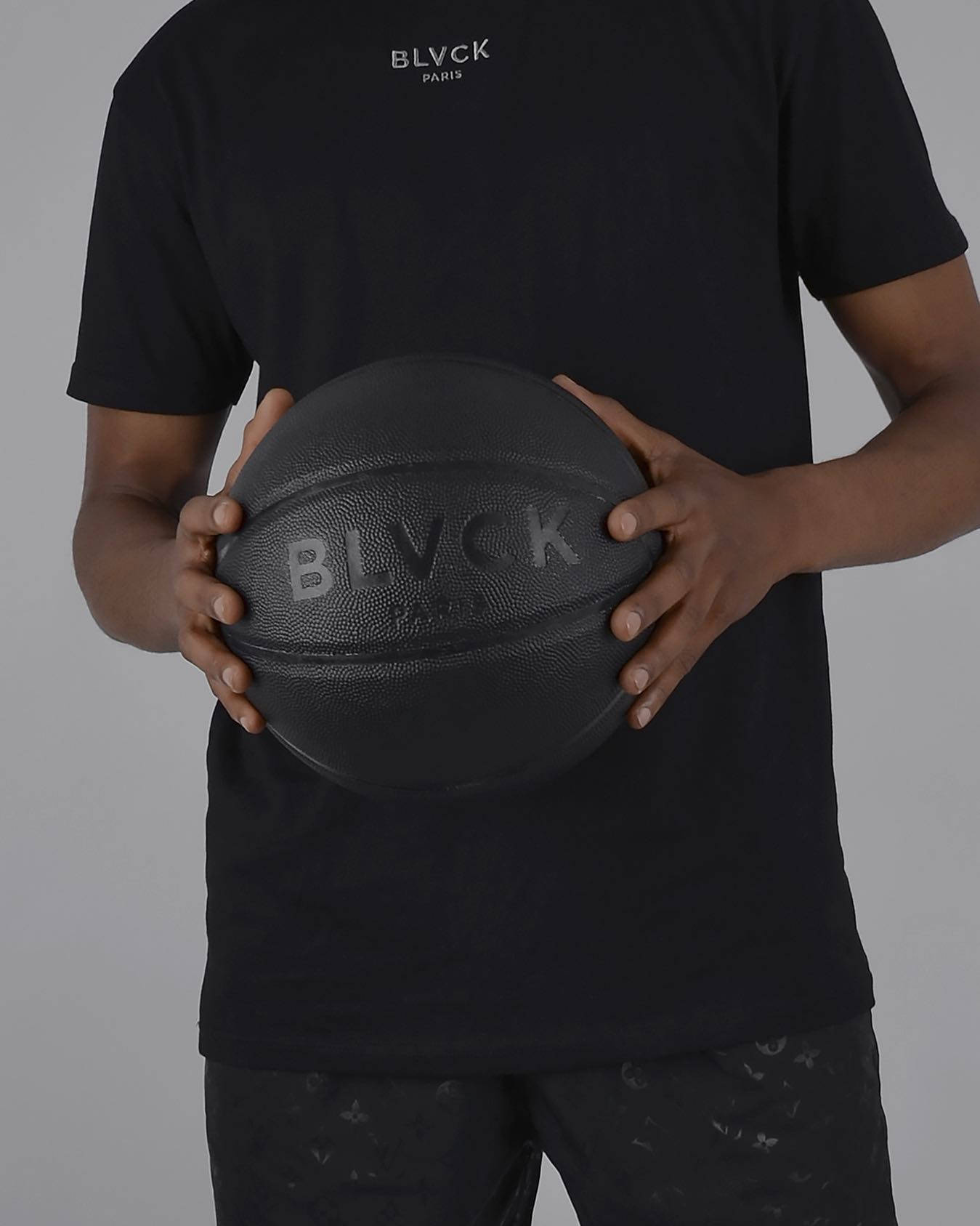 Blvck Paris 籃球商品 壁紙 — 一名身穿黑色服裝的人士，在純灰色背景前手持一個帶有壓印品牌標誌的啞光黑色籃球。