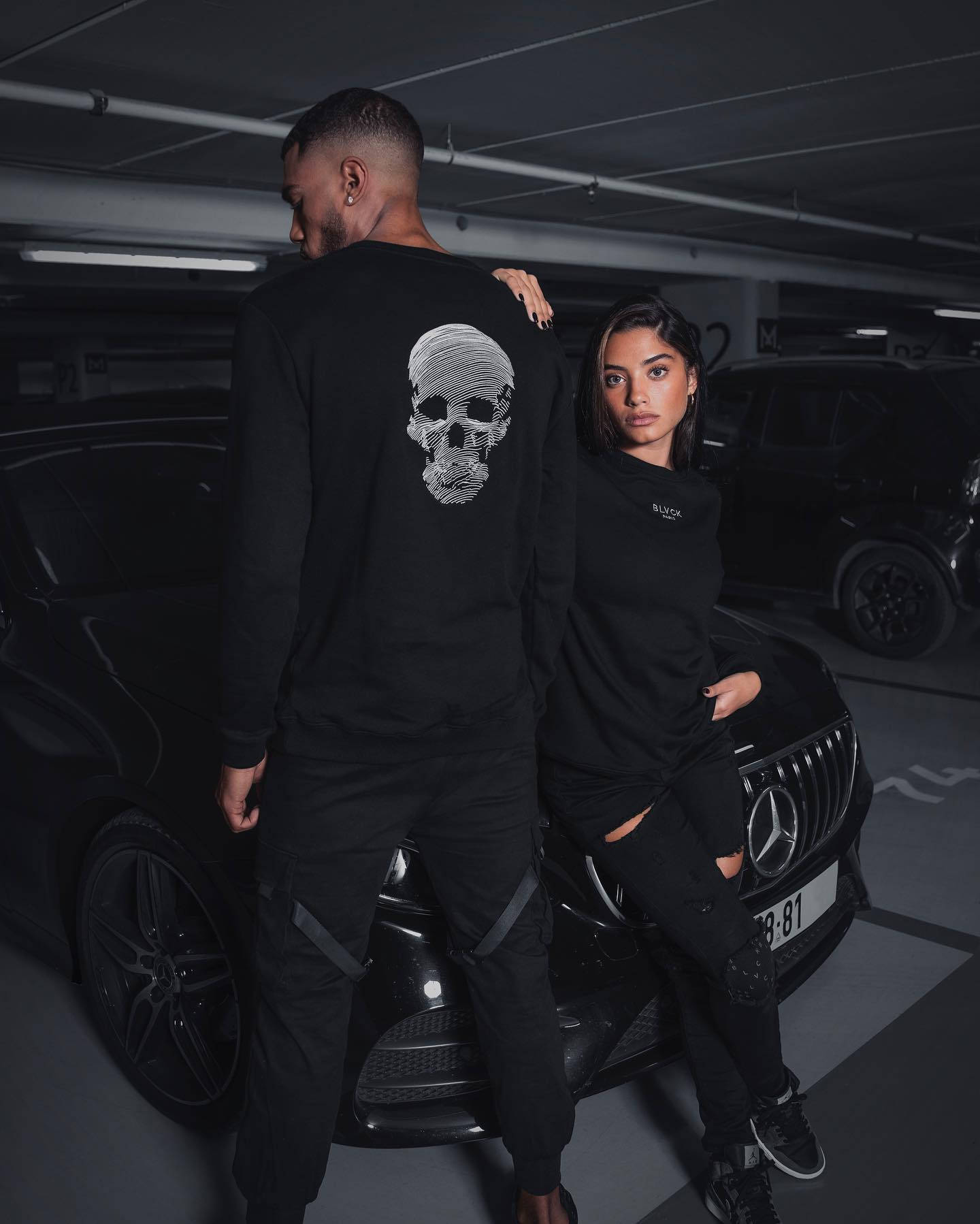 Blvck Paris 情侶穿搭 壁紙 — 一對穿著黑色服裝的情侶在光線昏暗的地下停車場，親密地站在一輛時尚的黑色豪華轎車旁。