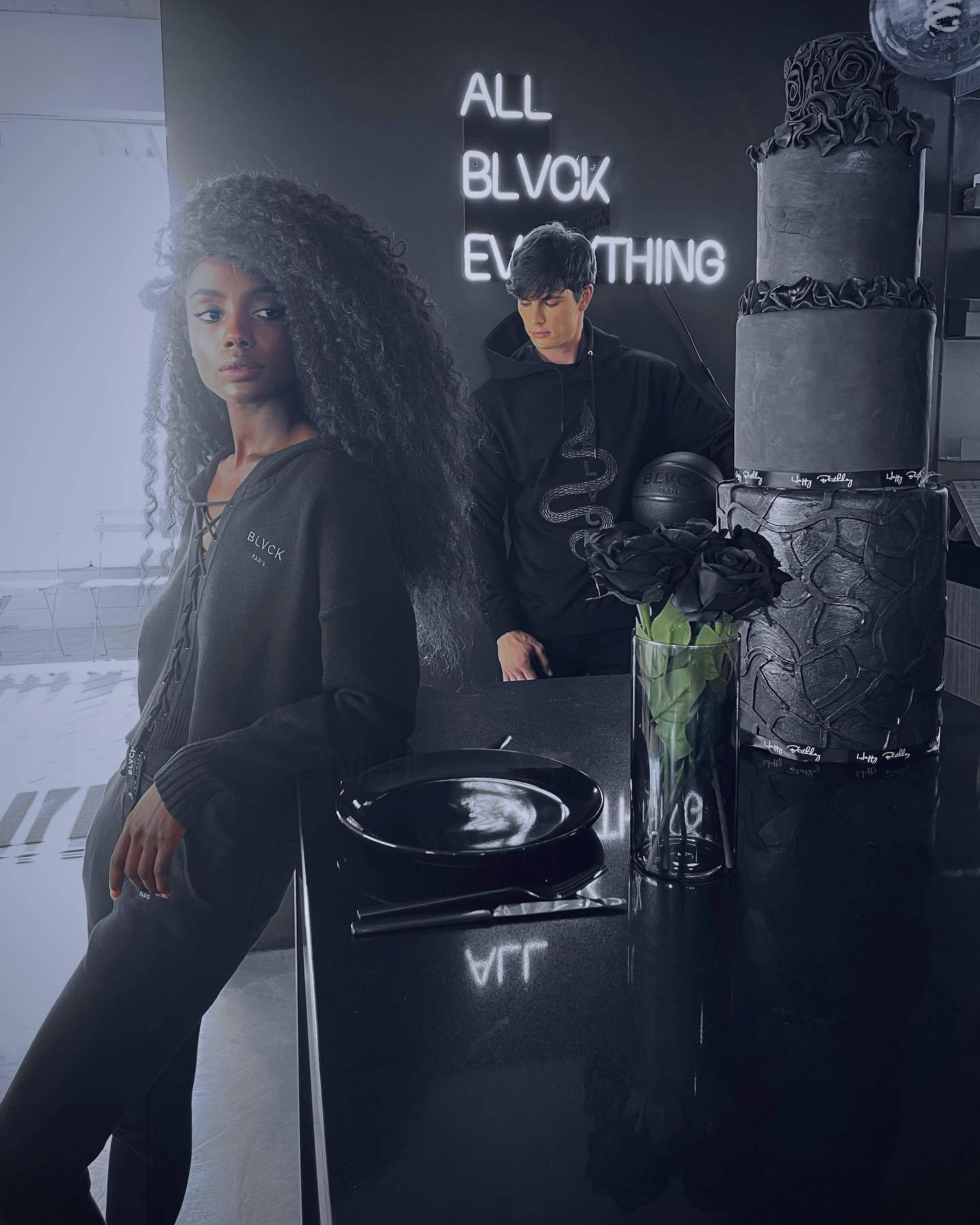 穿上 Blvck 風格，漫步巴黎街頭。 壁紙 — 兩個人在單色房間內，有霓虹燈牌和一個黑色蛋糕放在光面桌上。