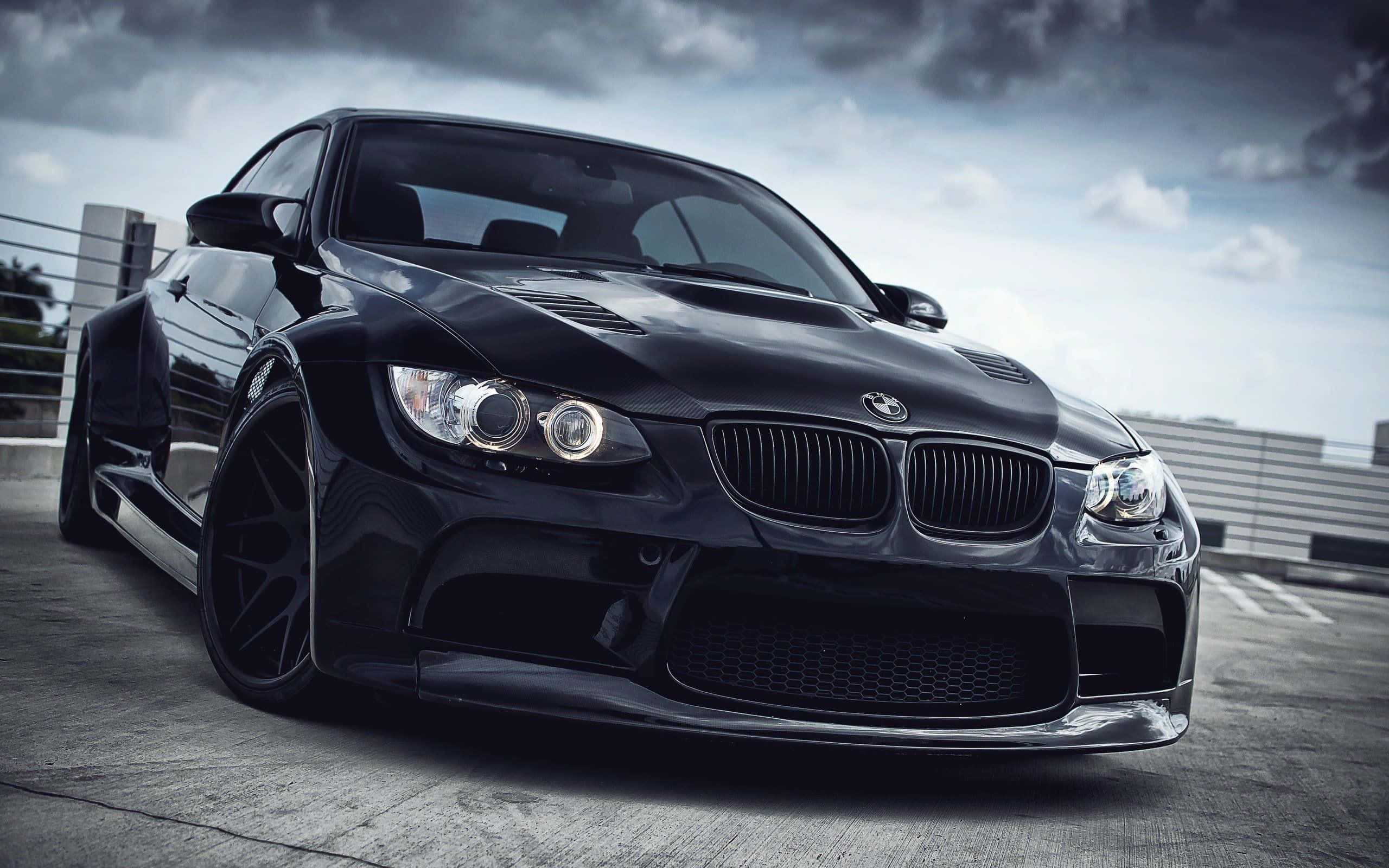 酷炫BMW 1系列汽車 壁紙 — 一輛時尚的黑色BMW M3跑車，配備定制寬體套件和發光頭燈，停在混凝土屋頂上，背景是戲劇性的多雲天空。