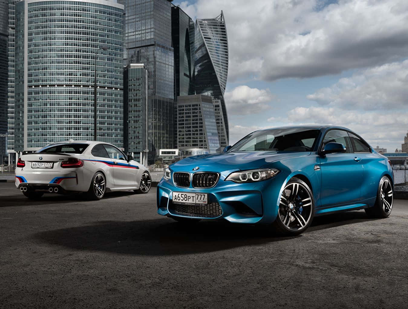 高畫質 BMW 平板壁紙 壁紙 — 兩輛時尚的BMW跑車，一藍一白，停在瀝青路面上，背景是現代玻璃摩天大樓，天空多雲。