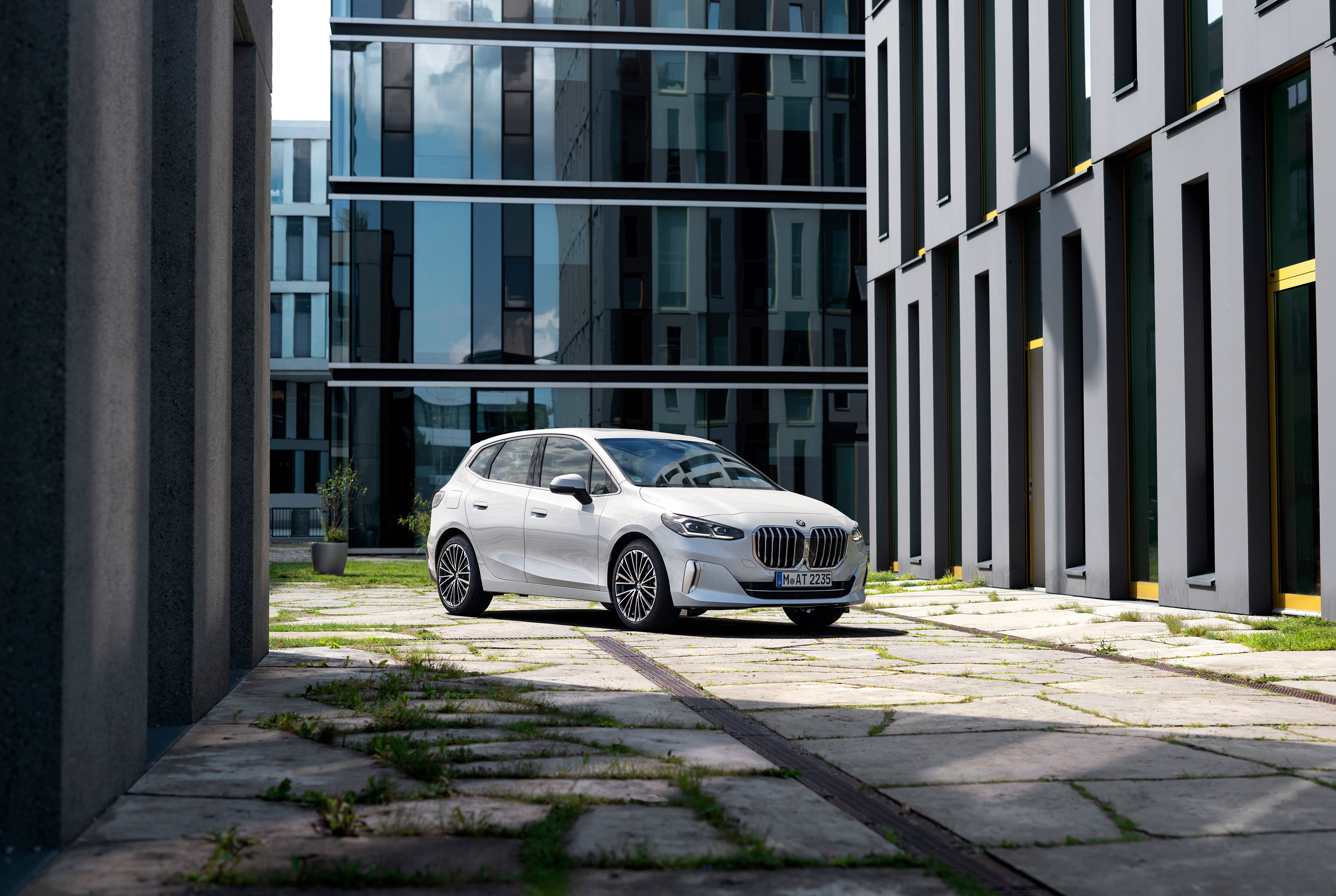 BMW 223i 豪華汽車轉彎 壁紙 — 一架時尚白色BMW掀背車停泊在現代玻璃與灰色混凝土建築之間，石鋪地上長出青草。