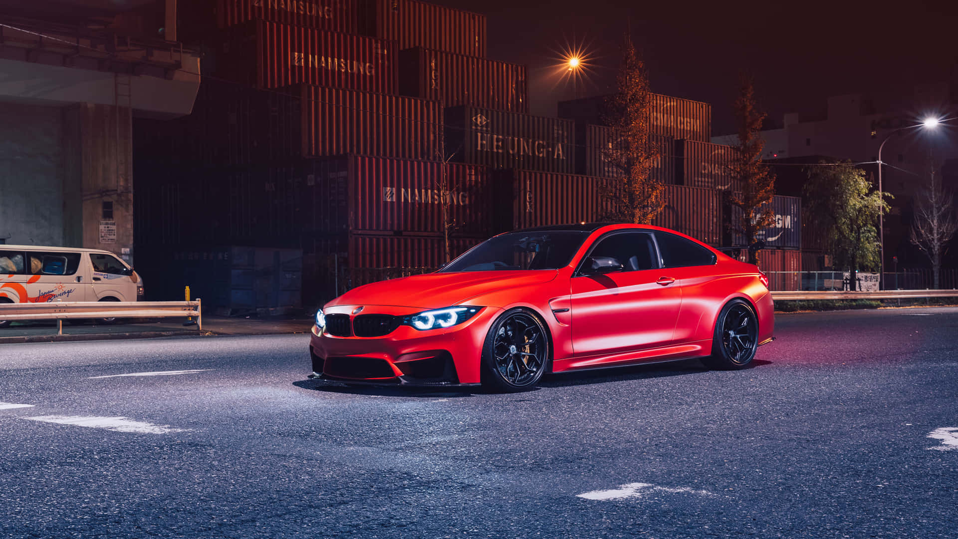 夜間濕潤的城市街道上停放的紅色BMW M4，車頭燈閃爍，後方有堆疊的貨運集裝箱。