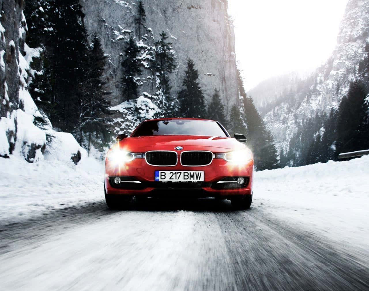 BMW 320d 紅色雪地行駛平板壁紙 壁紙 — 一輛鮮紅色的BMW跑車在白雪覆蓋的山路上飛馳，兩旁是高聳的常綠樹木和岩石峭壁。