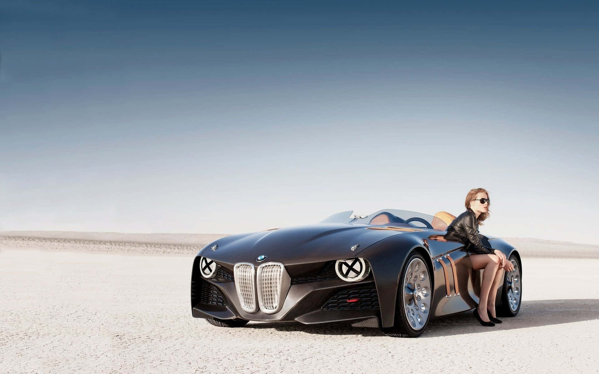 經典BMW 328 壁紙 — 一架時尚黑色概念車，一位戴太陽眼鏡嘅女士喺晴朗天空下嘅廣闊沙漠平原上挨喺車旁邊休息。
