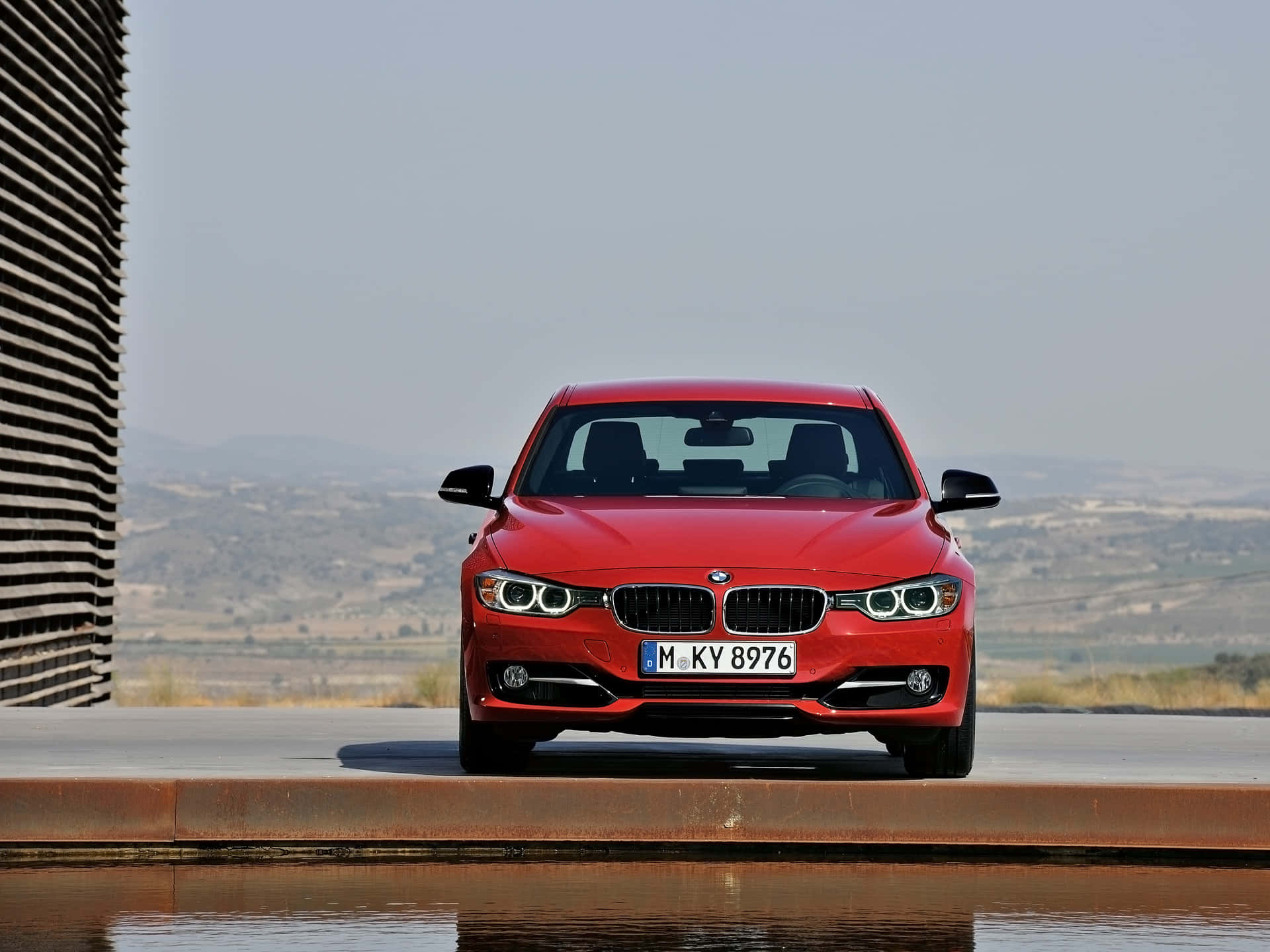 穿梭城市街頭的時尚BMW 328 壁紙 — 一輛鮮紅色的BMW房車在混凝土平台上向前行駛，旁邊有水，背景有現代條紋牆和朦朧的山丘。