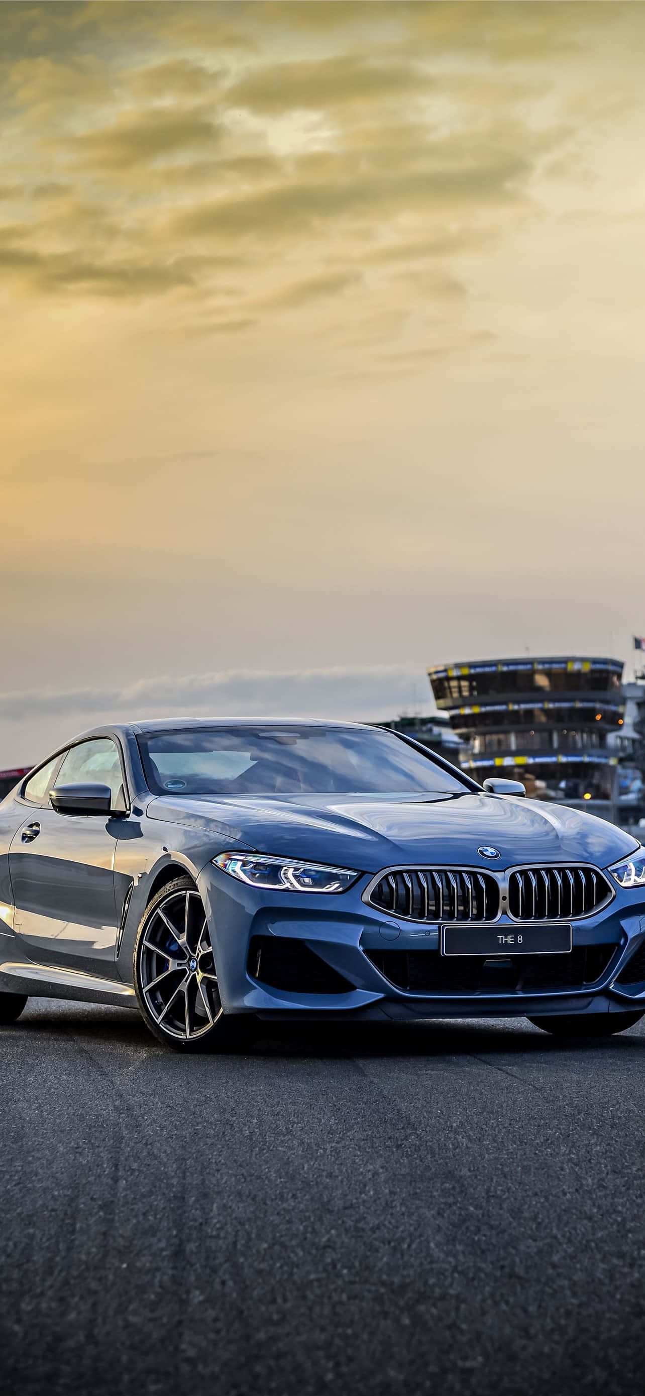BMW 328 的力量與美學 壁紙 — 一架時尚的藍色BMW跑車停泊在瀝青路上，背景是金色夕陽天空和賽車場看台。