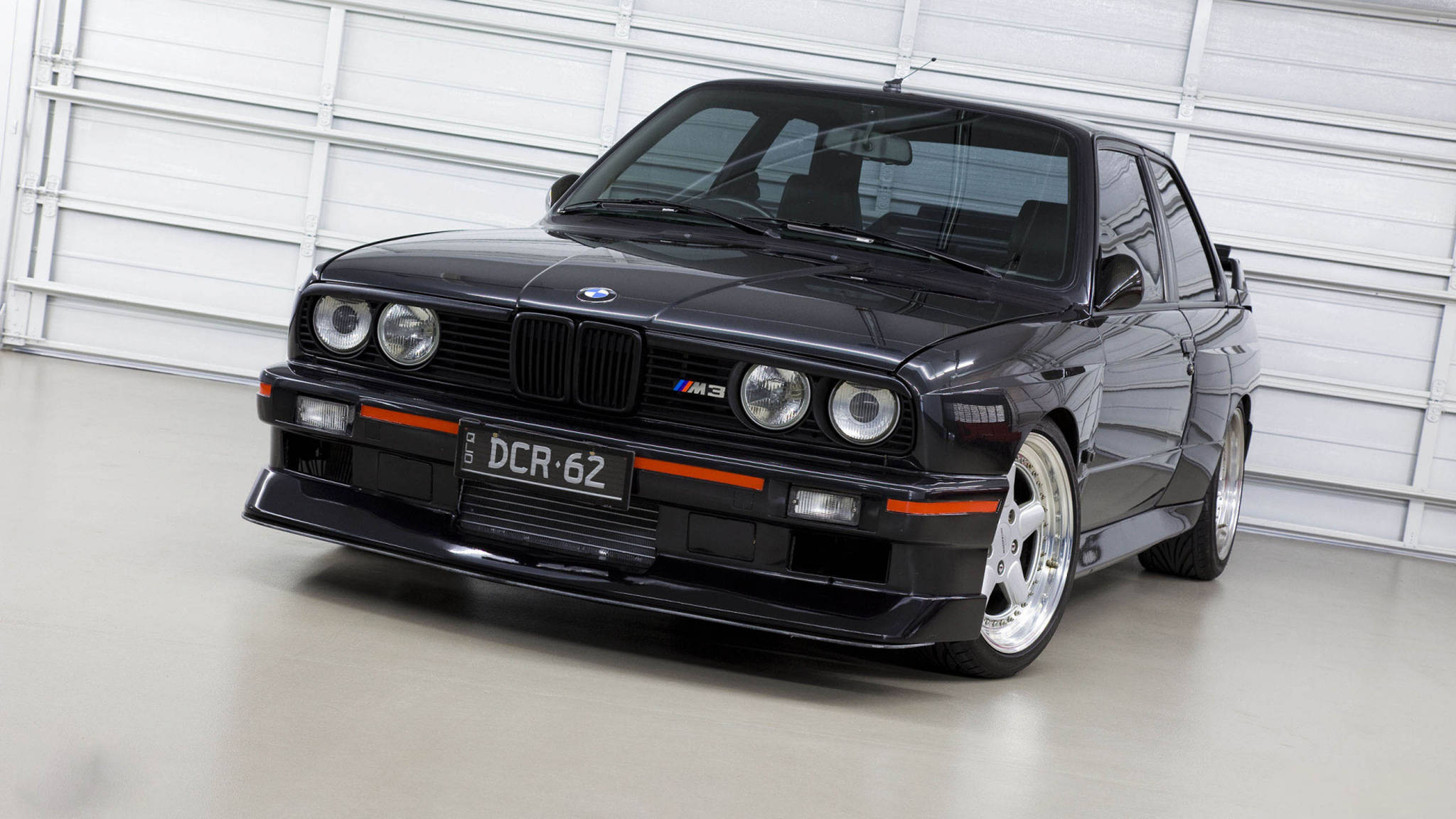 黑色經典BMW 3系列E30 壁紙 — 一輛時尚的黑色BMW M3跑車帶有橙色點綴，停在潔白的車庫中。