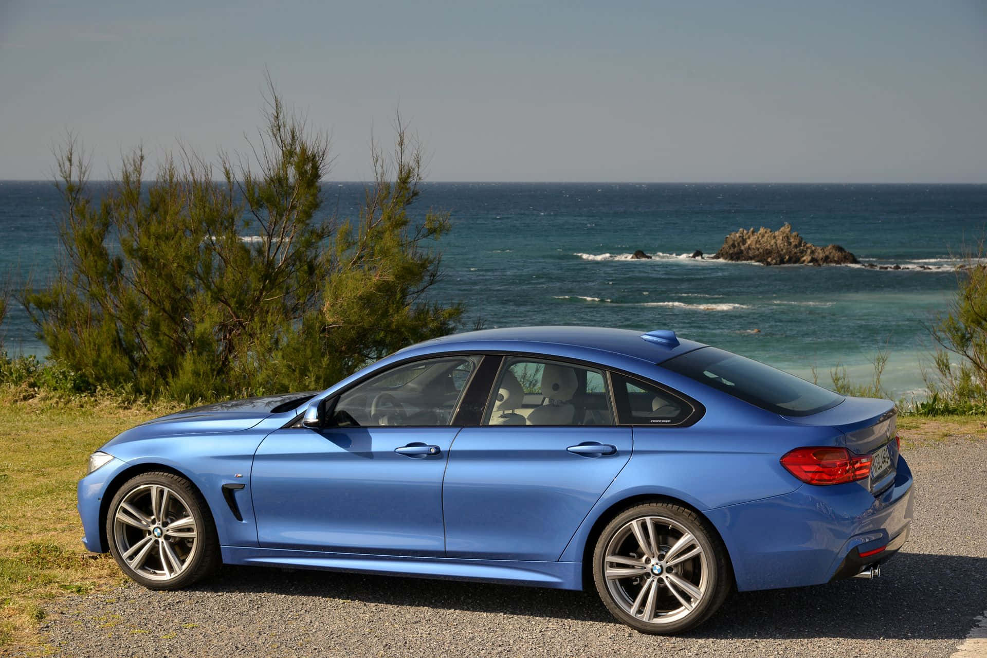 動感奔馳 BMW 4 系列 壁紙 — 一架時尚的藍色寶馬轎跑車停泊在海邊的碎石路上，背景有綠色灌木和碧綠的海水。