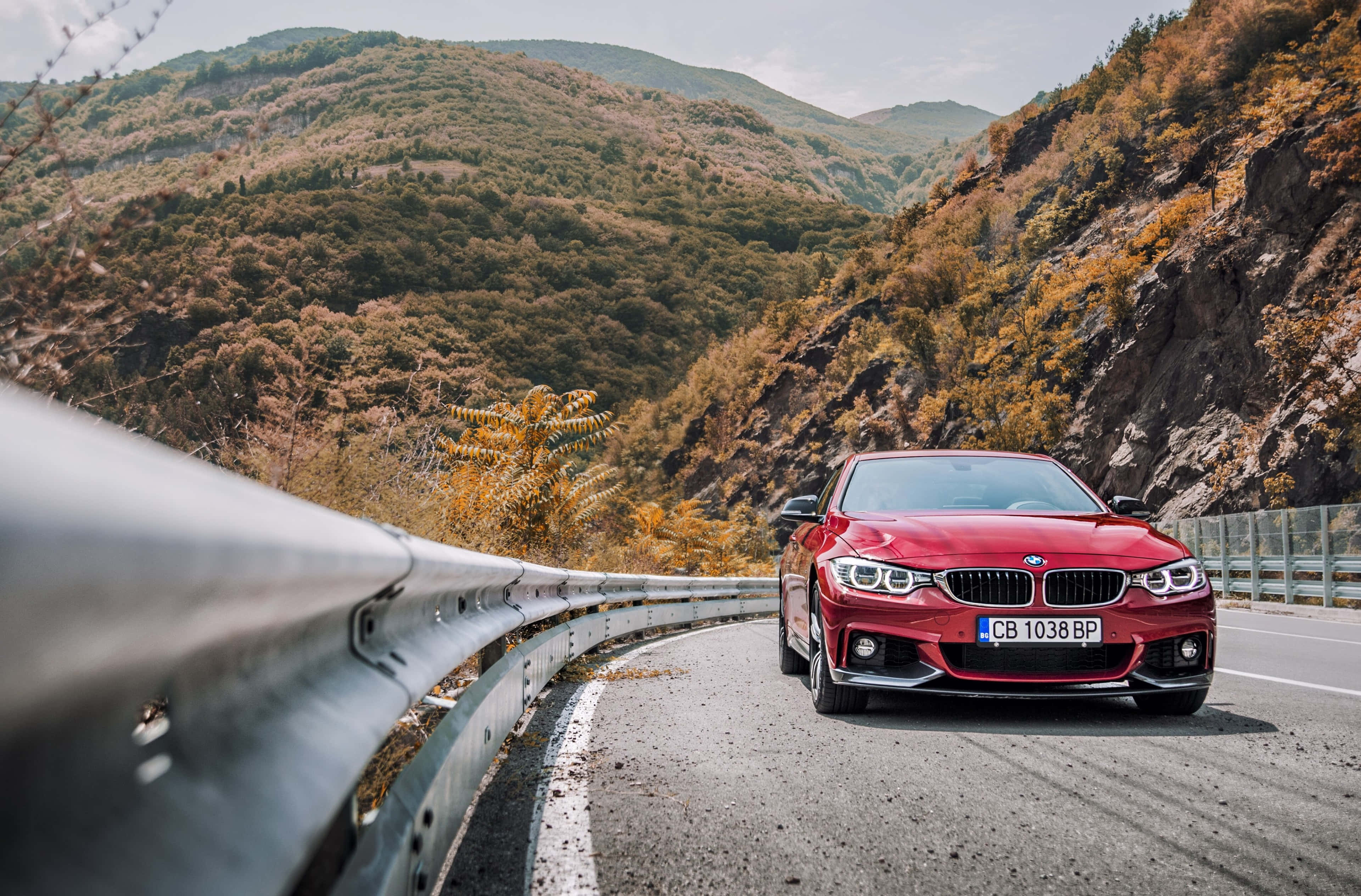 一款大膽的 BMW 440i 壁紙 — 一輛鮮紅色的BMW跑車沿著蜿蜒的山路行駛，兩旁是秋色的山丘和護欄。