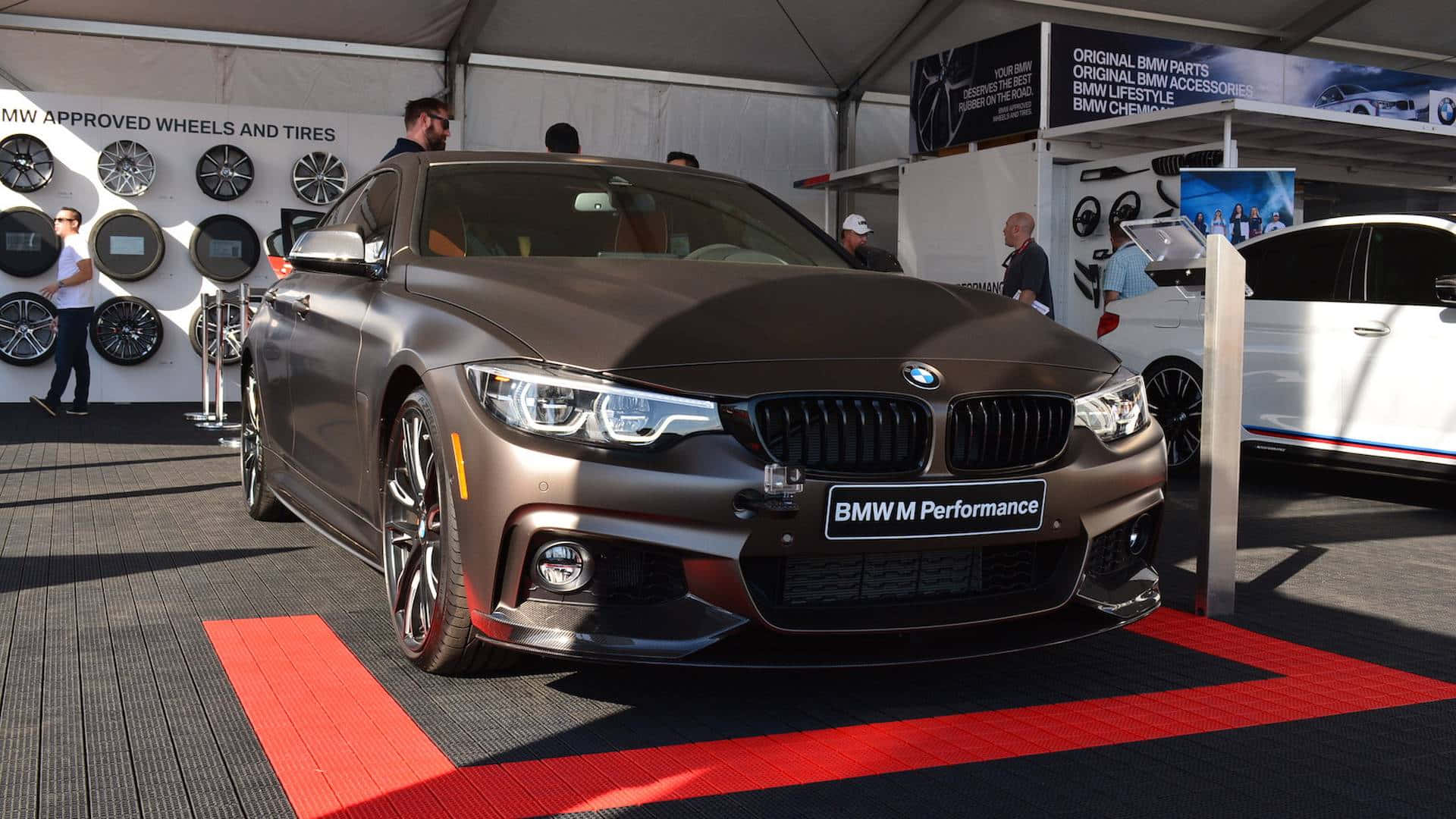 啞光棕色BMW M4雙門轎跑車在車展紅地毯上，背景有輪胎展示。