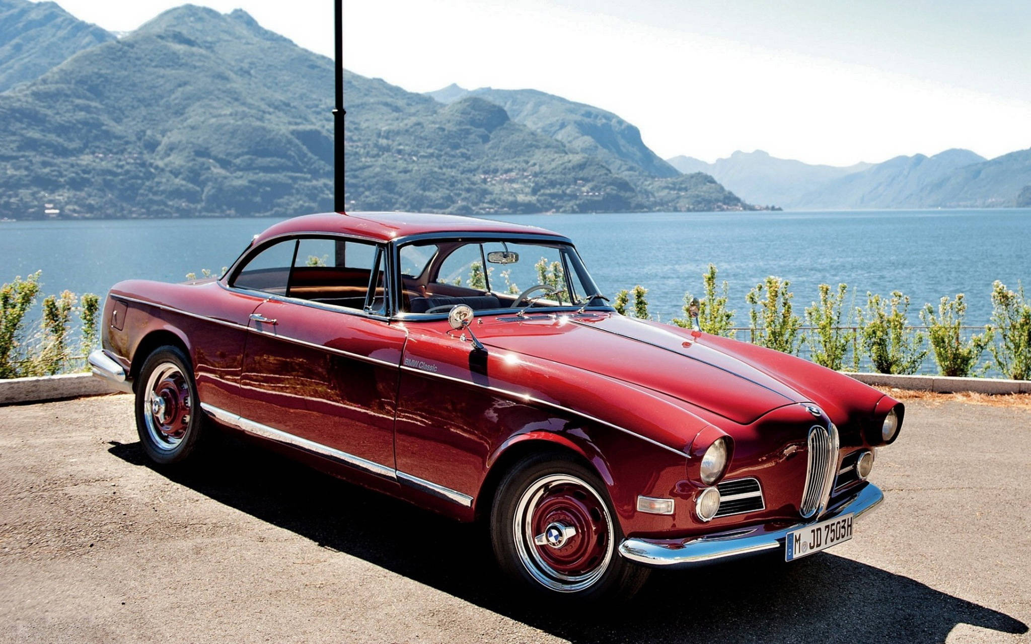 紅色經典BMW 506 壁紙 — 一輛光澤的紅色復古跑車停泊在寧靜的湖邊，背景是山巒，在明亮的日光下。