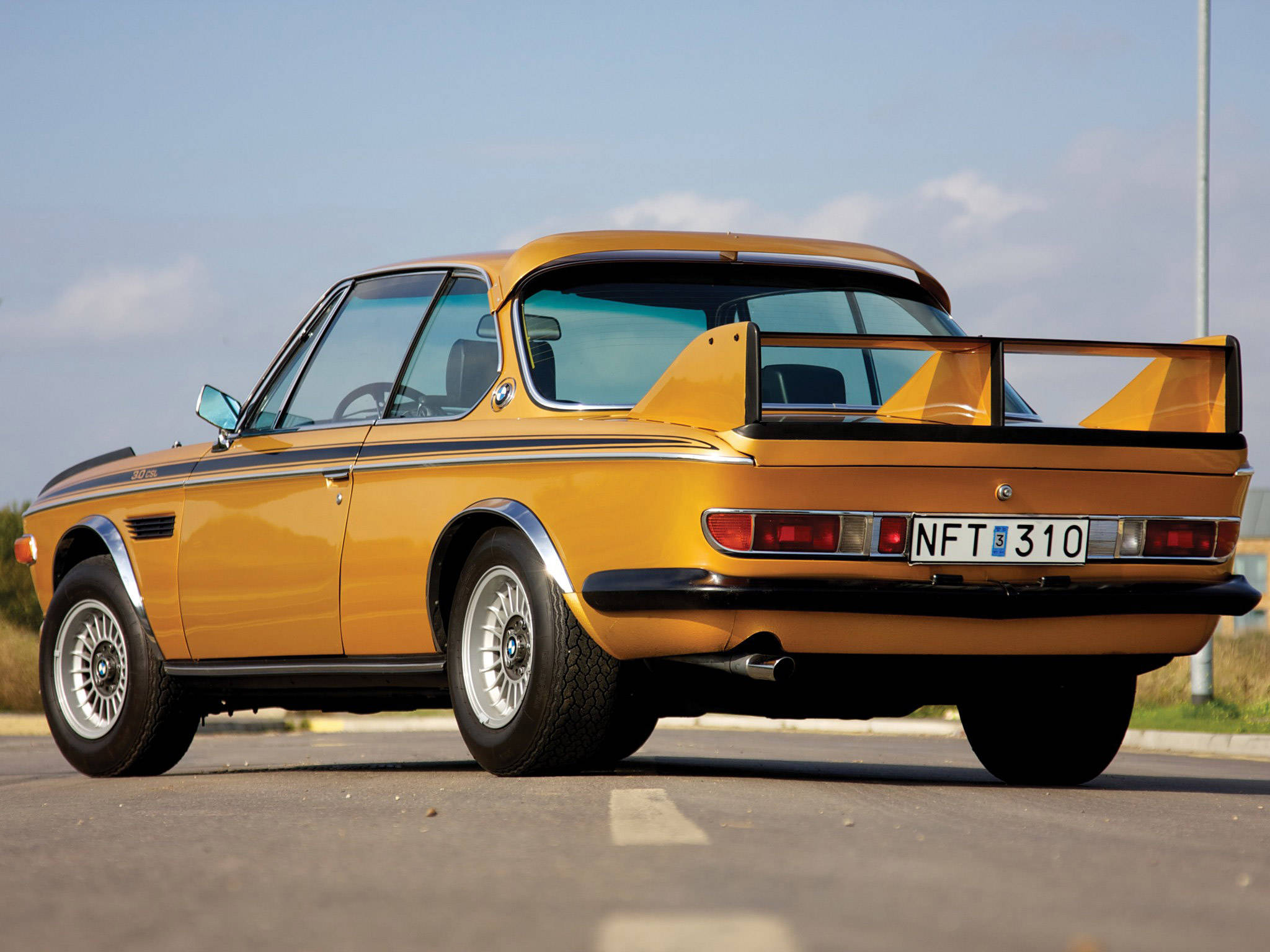 金屬金色經典BMW CSL-E9 壁紙 — 一輛充滿活力的黃色經典BMW 3.0 CSL跑車，帶有大型後擾流板，停在晴朗藍天下的瀝青路上。