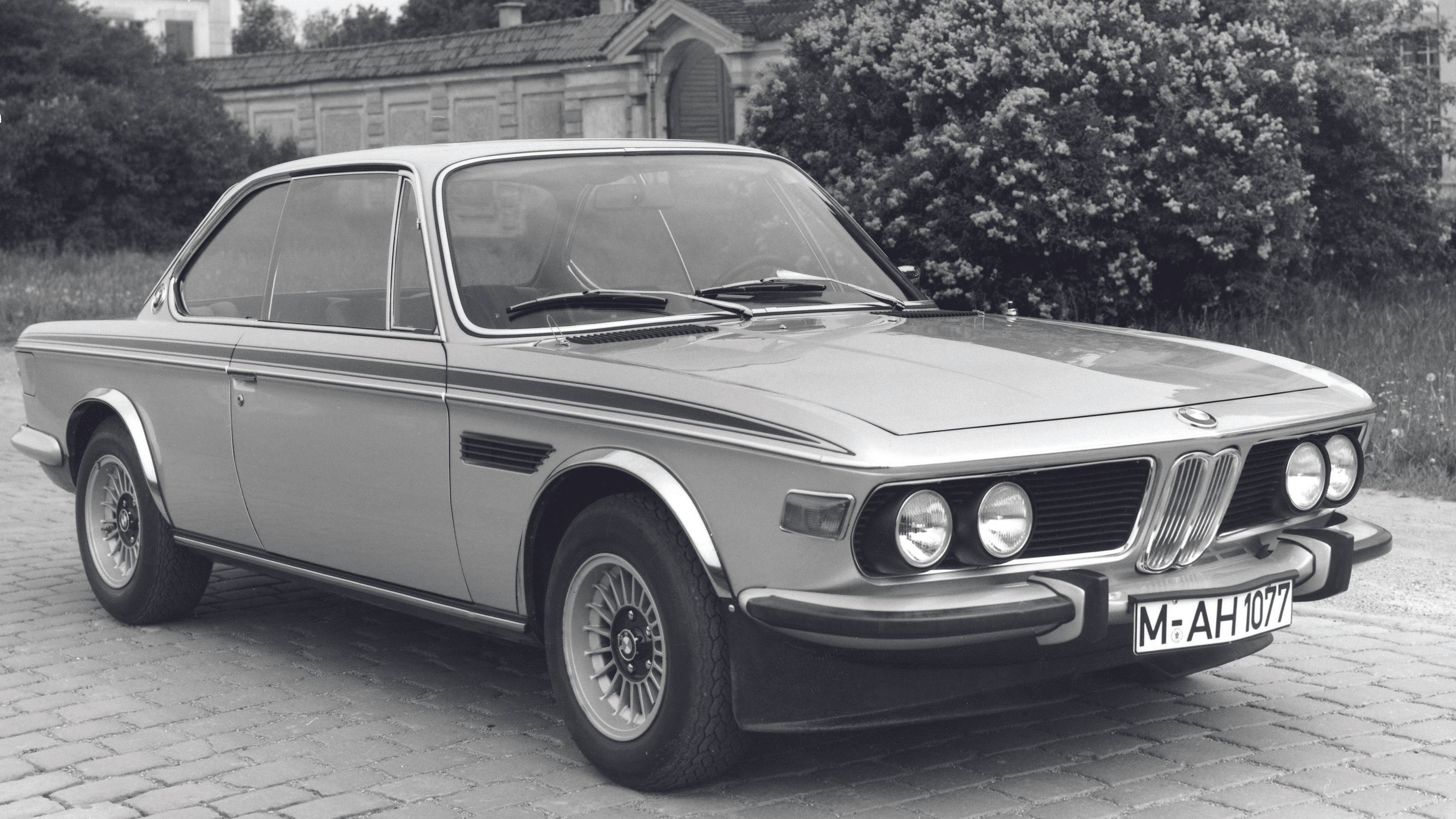 灰階經典BMW E9 壁紙 — 一輛經典銀色寶馬雙門轎跑車停泊在鵝卵石車道上，背景有樹木和建築物。