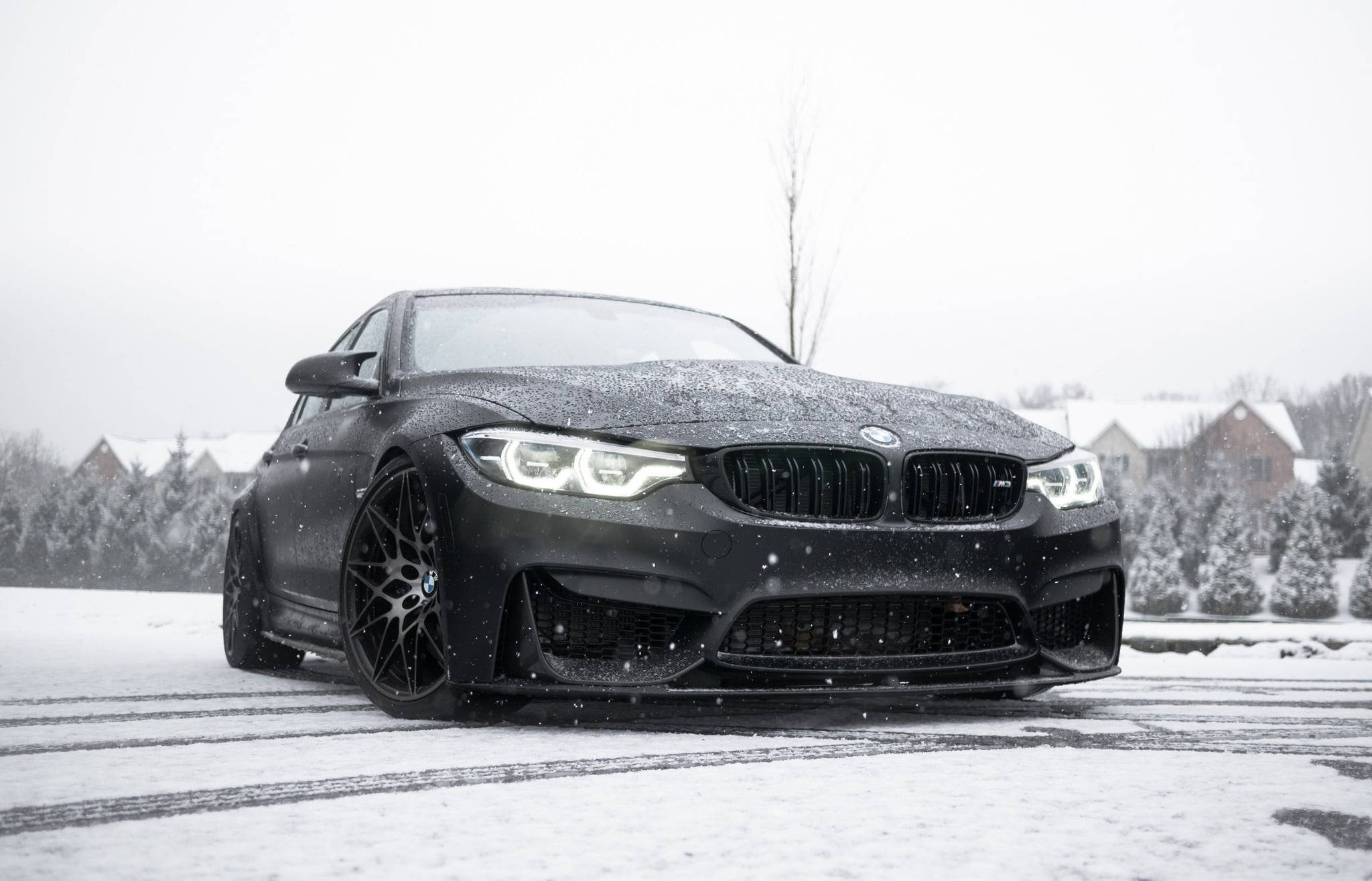 雪覆霧面黑 BMW 電腦壁紙 壁紙 — 一輛啞光黑色BMW M4在郊區街道上飄移穿過飄落的雪花，背景是積雪的房屋和樹木。