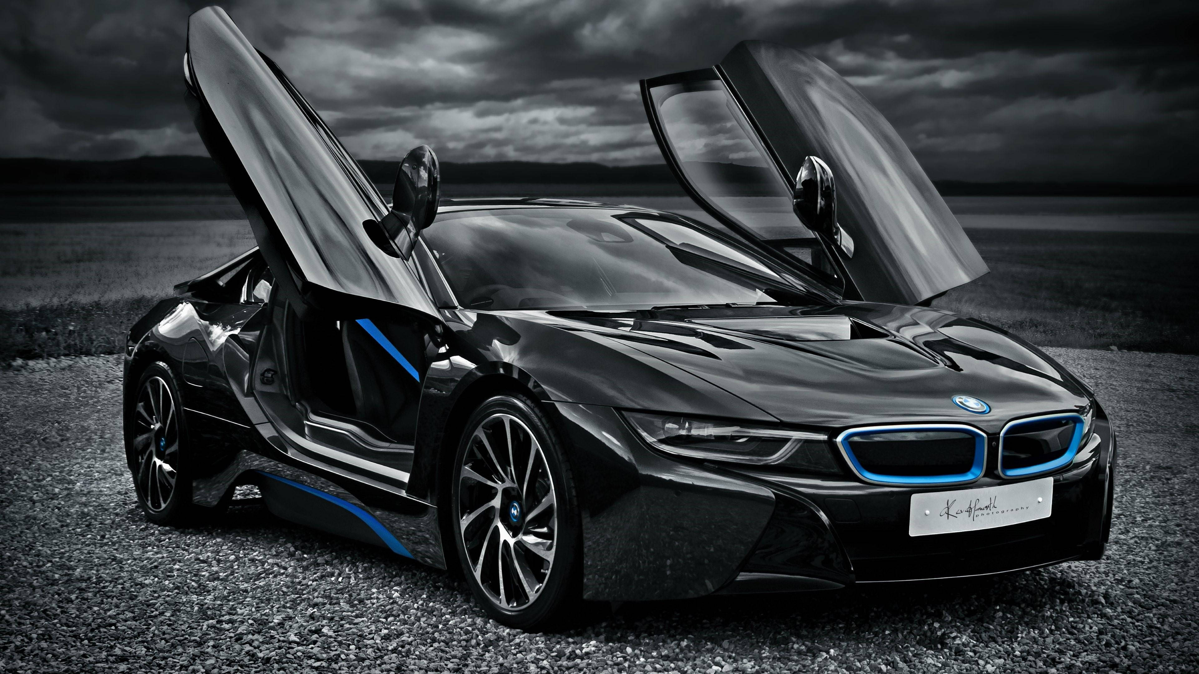 酷炫黑色 BMW i8 電腦壁紙 壁紙 — 一架時尚的黑色BMW i8跑車，鷗翼車門打開，停在碎石上，背景是風暴天空，帶有藍色發光點綴。